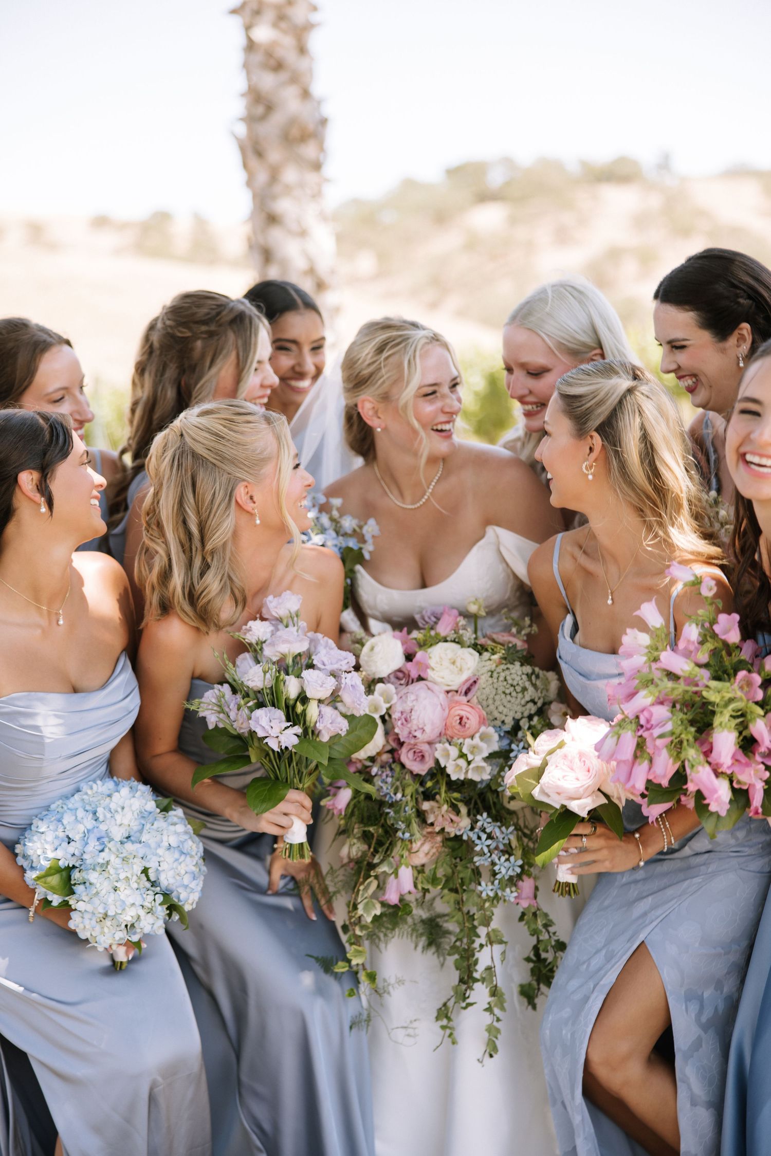 Bride laughing with bridesmaids holding pink and blue floral bouquets outdoors.
