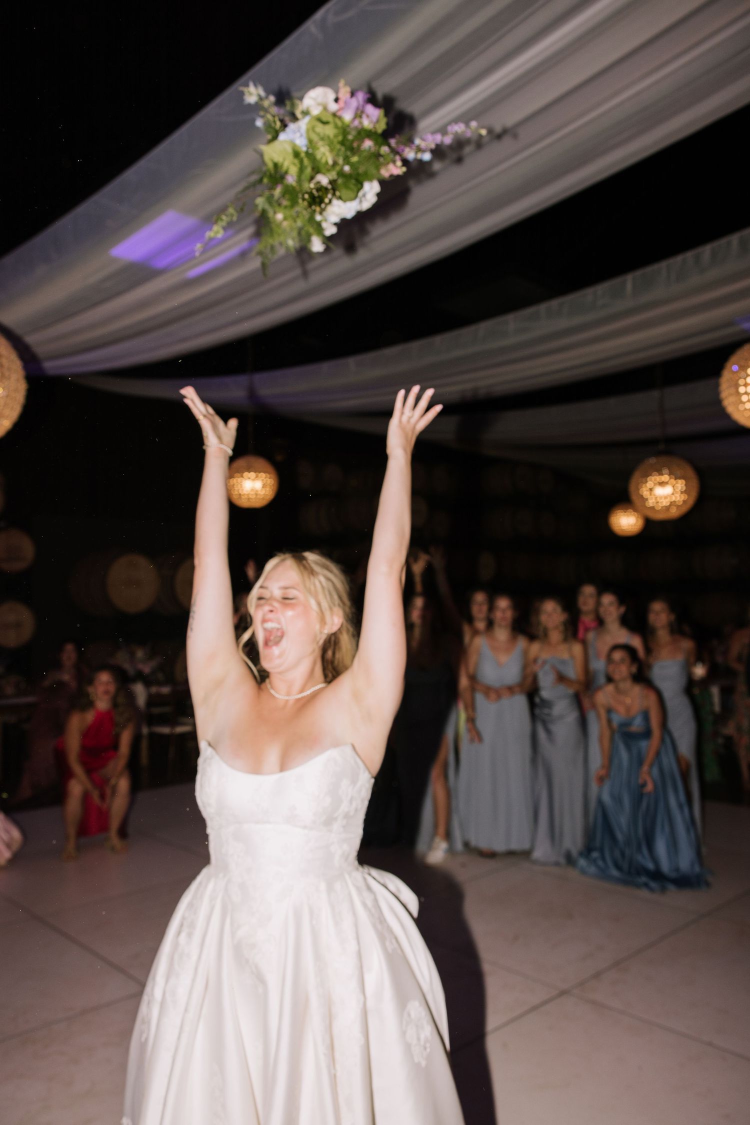 Bride in white ball gown tosses bouquet of purple and green flowers at indoor wedding reception.
