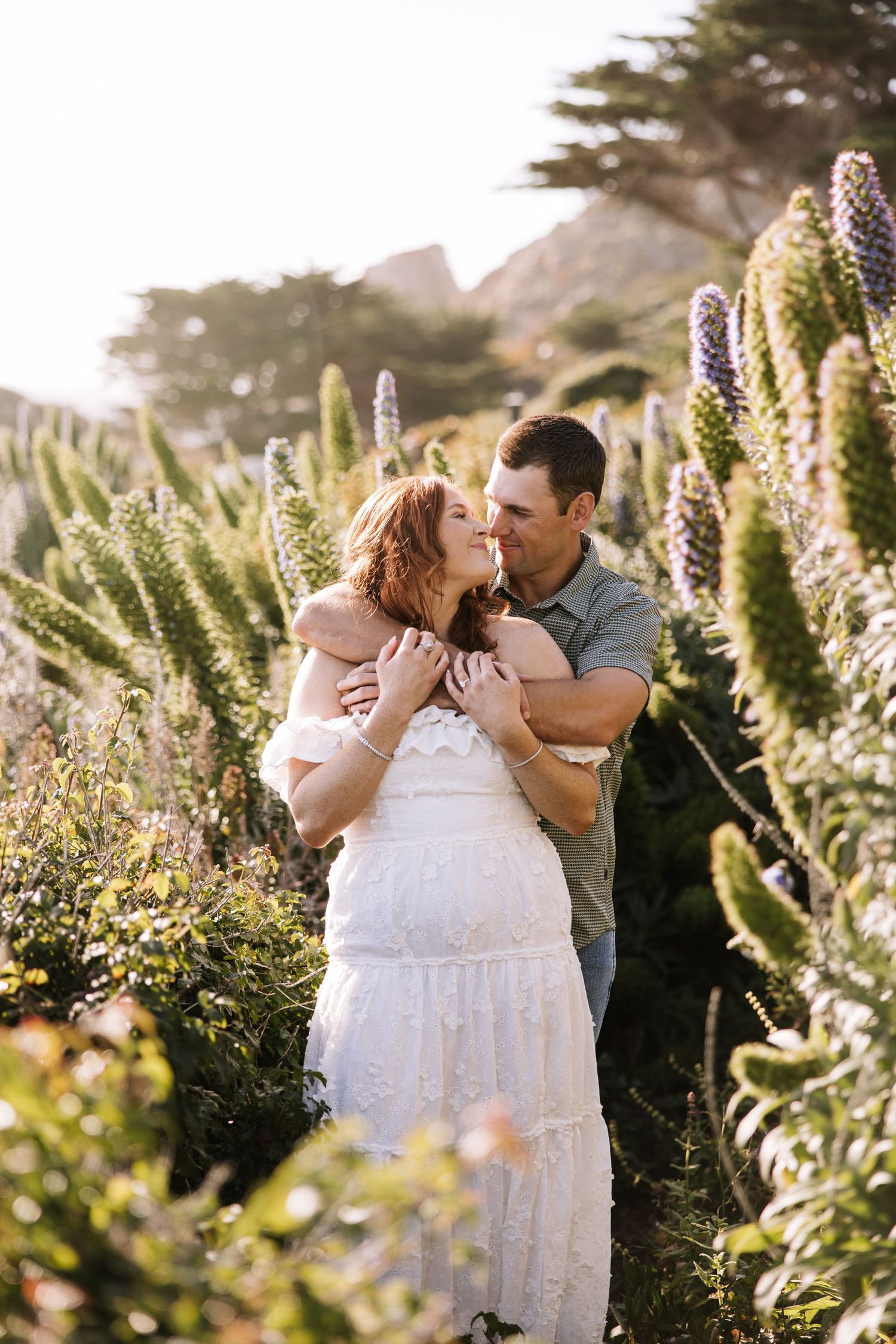 Couple sharing a kiss surrounded by tall purple wildflowers in golden coastal landscape.