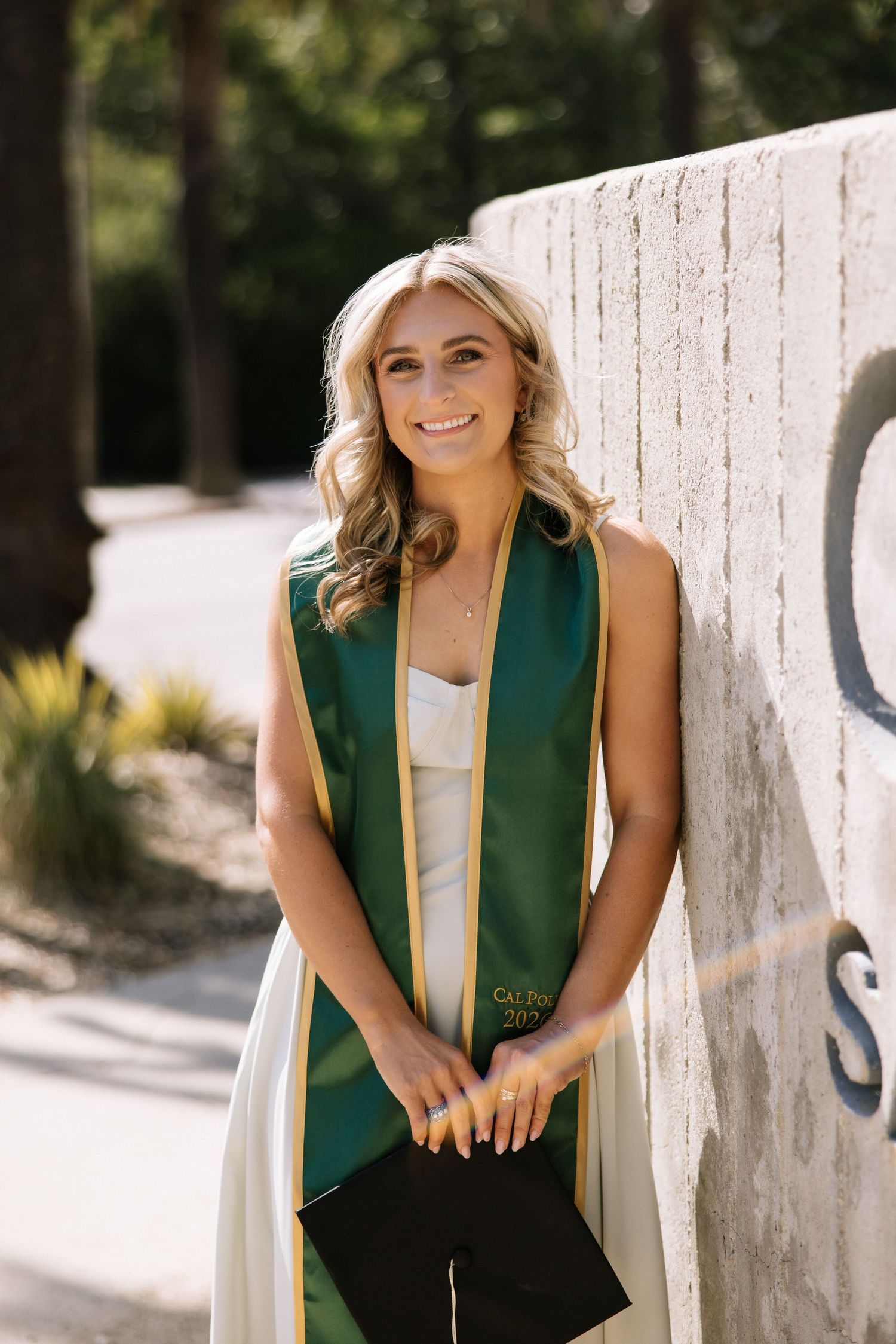 Smiling blonde woman in a white dress wearing a green and gold Cal Poly graduation stole leaning against a wall.