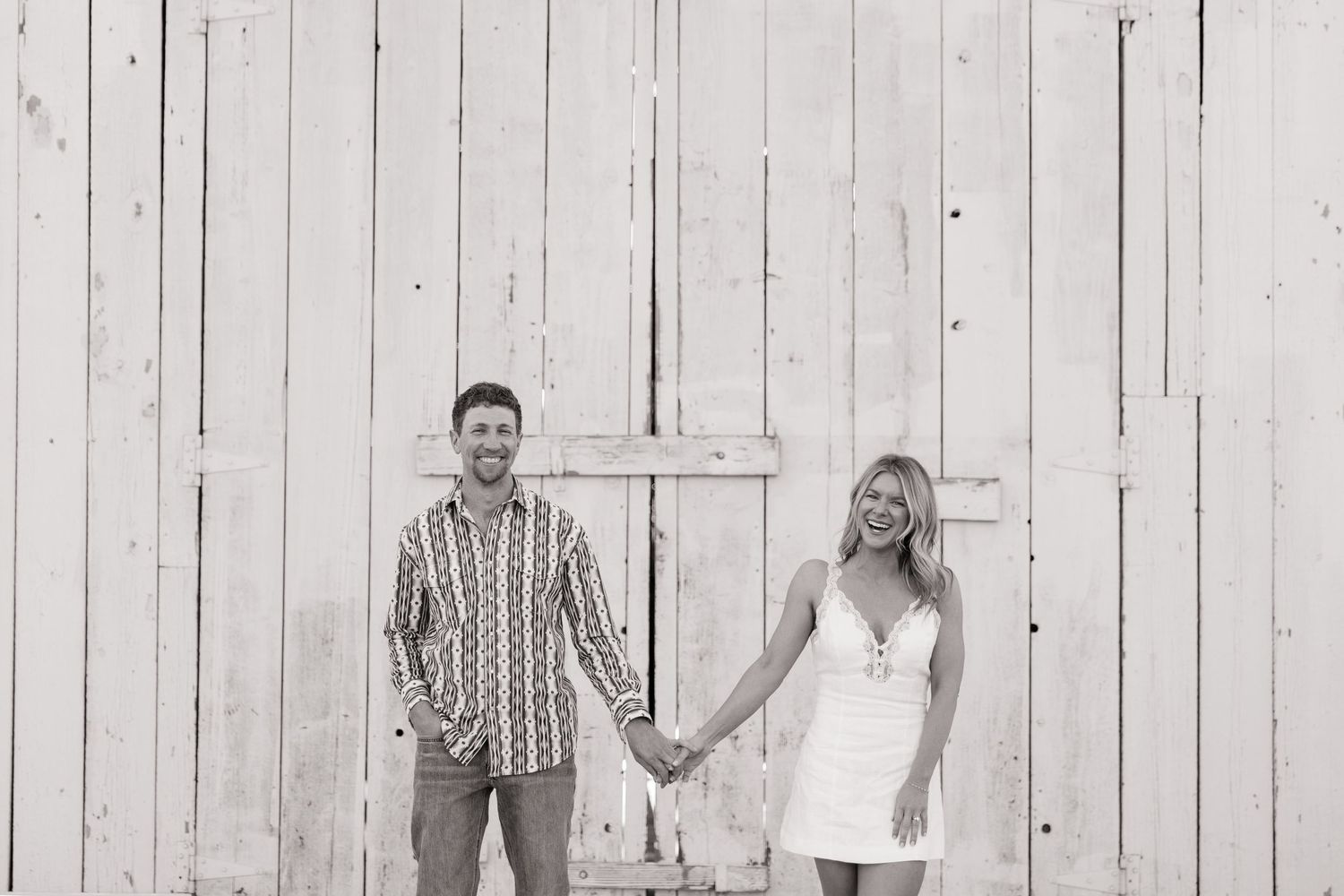 Black and white photo of a smiling couple holding hands in front of a white barn door.