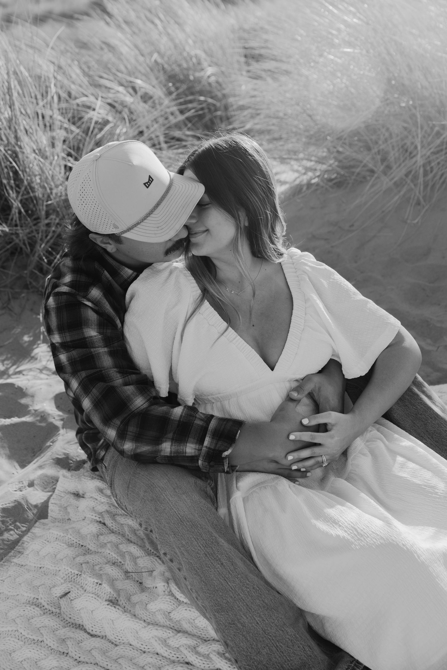 Black and white photo of couple sitting in sand dunes, man in plaid shirt embracing pregnant partner.