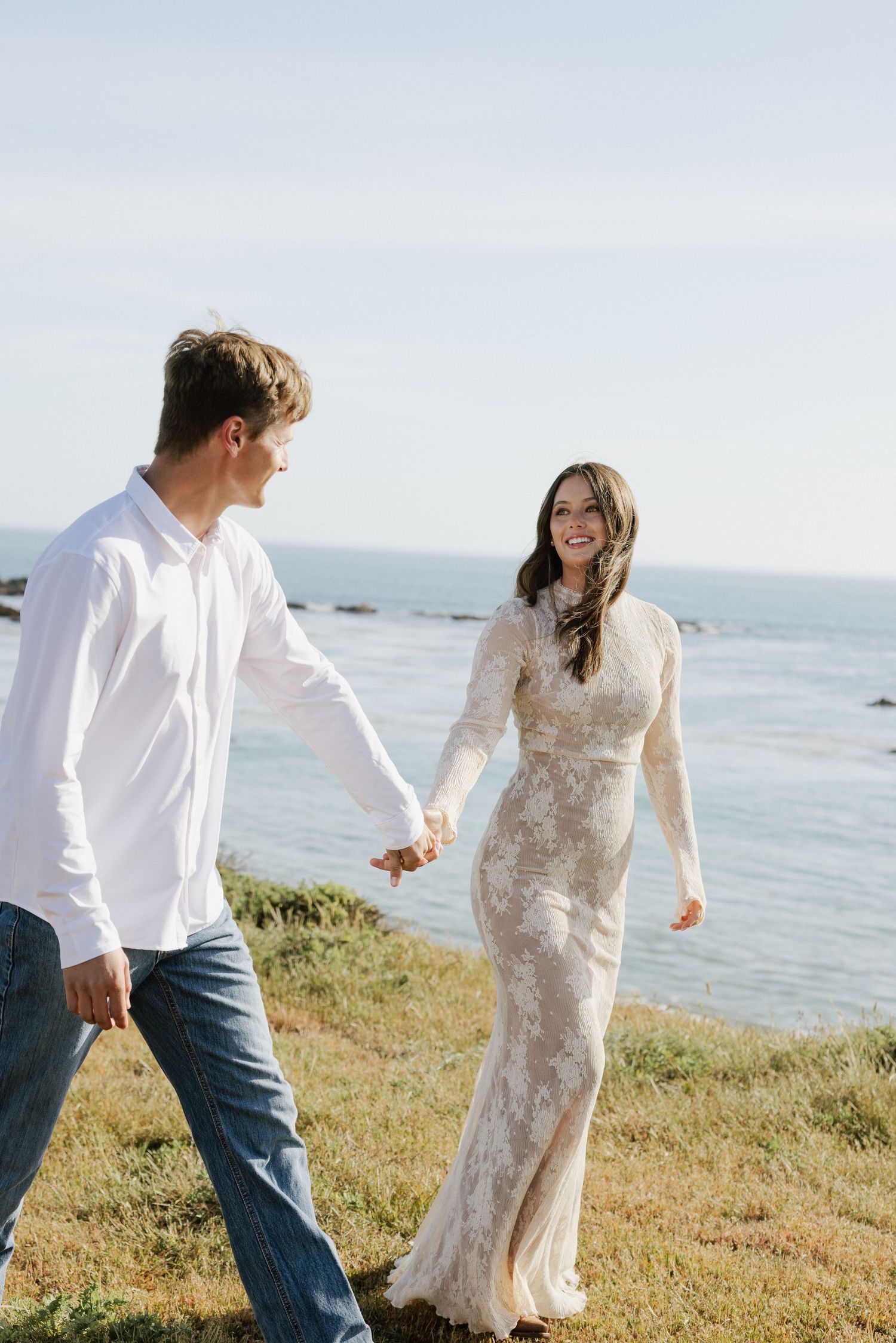 Smiling woman in lace wedding dress holding hands with man in white shirt walking by ocean.