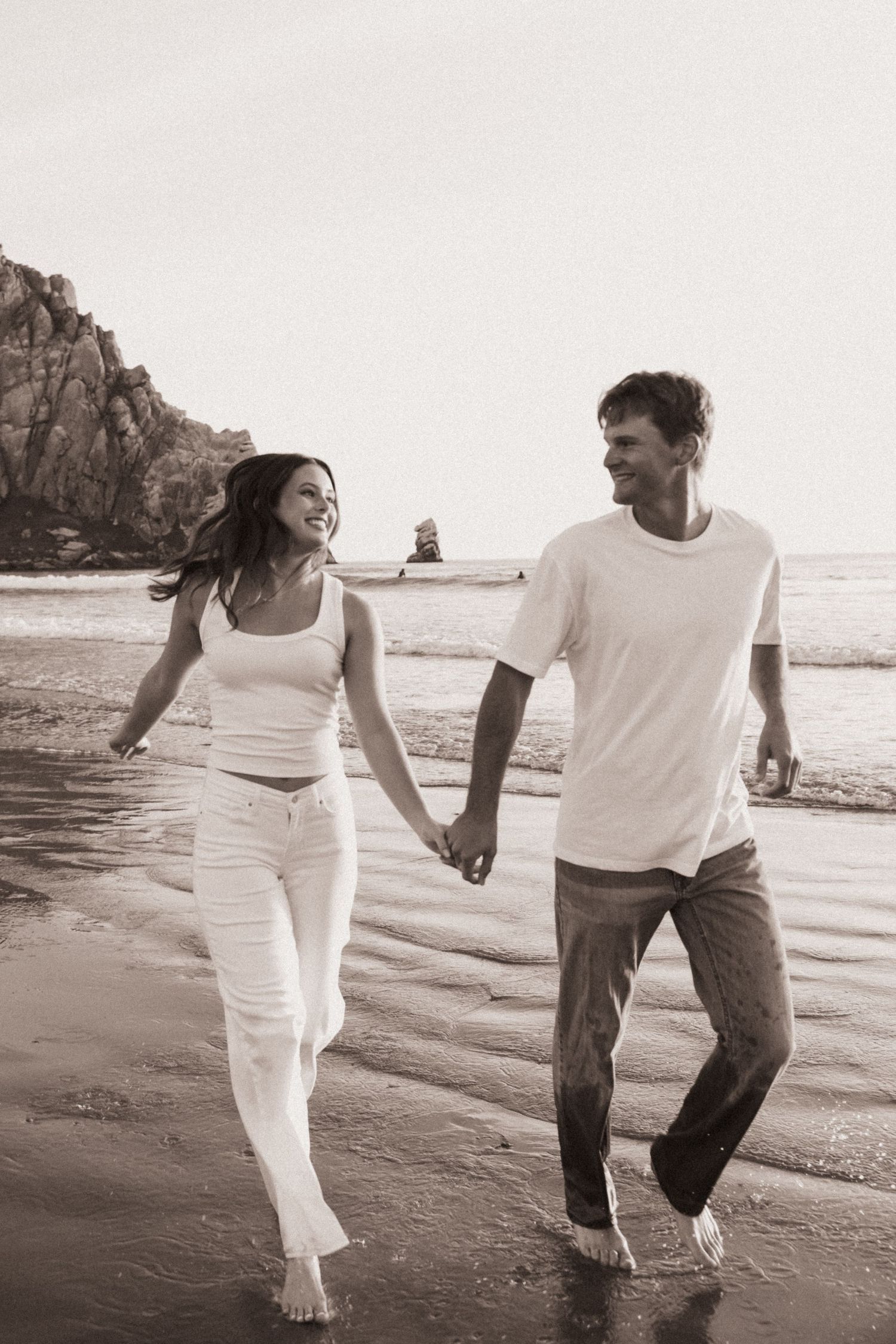 Happy couple holding hands and walking barefoot on beach in black and white photo.