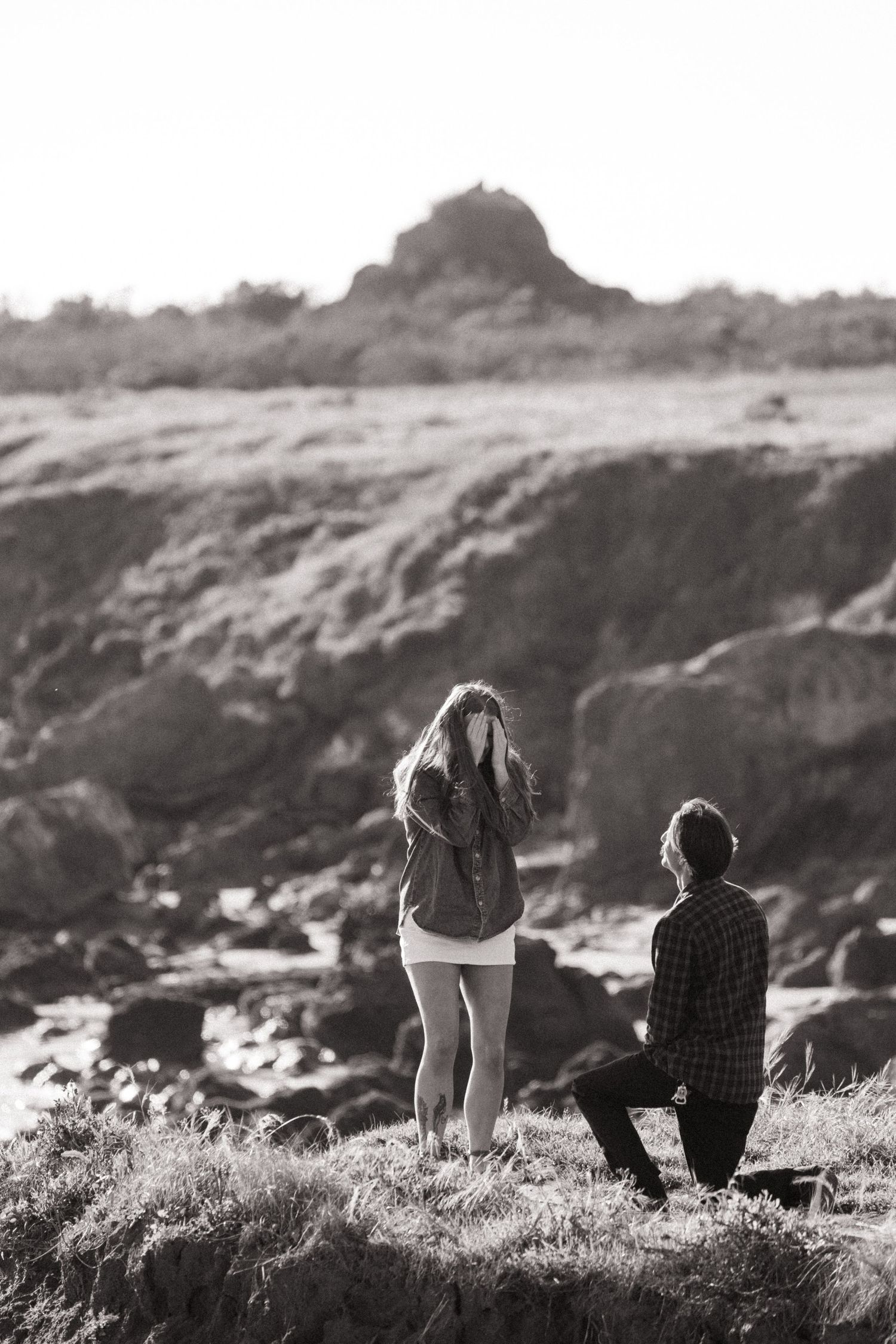 Black and white photo of a man proposing on one knee to a surprised woman on rocky coastal cliffs.
