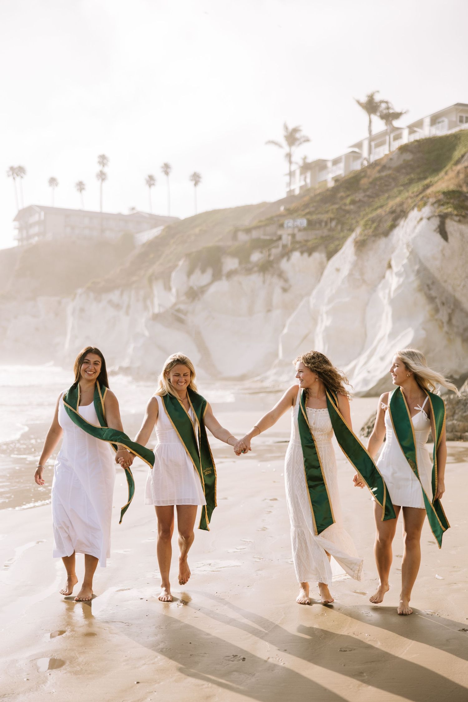 Four joyful female graduates in white dresses and green sashes walk hand in hand on sunny beach.