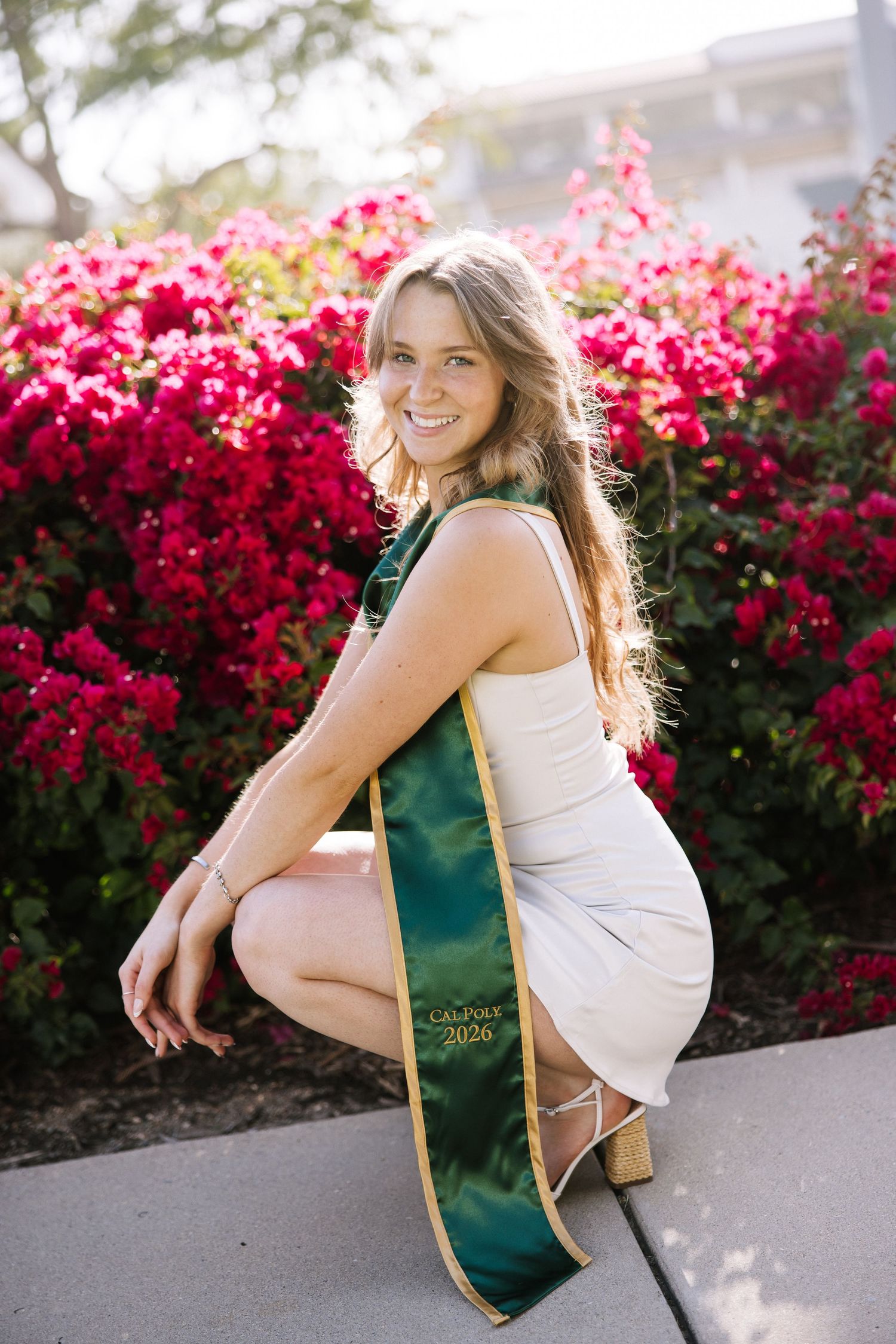 Graduate in white dress crouches with green Cal Poly sash before vibrant red flowers.