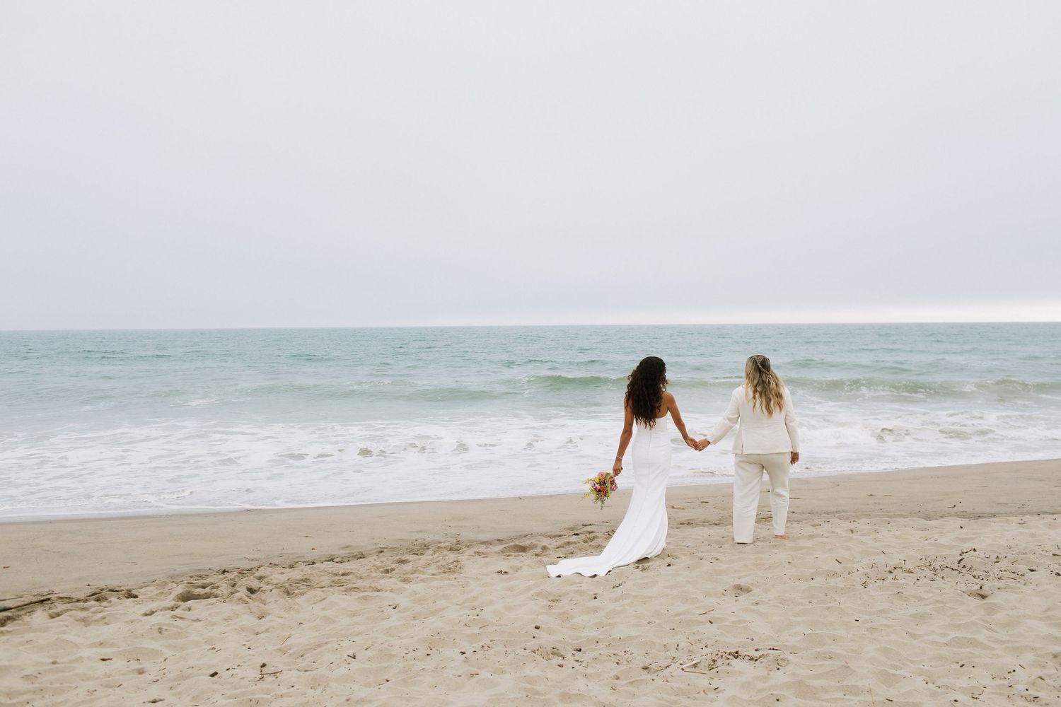 Two brides in white hold hands on a sandy beach, facing the ocean waves on their wedding day.