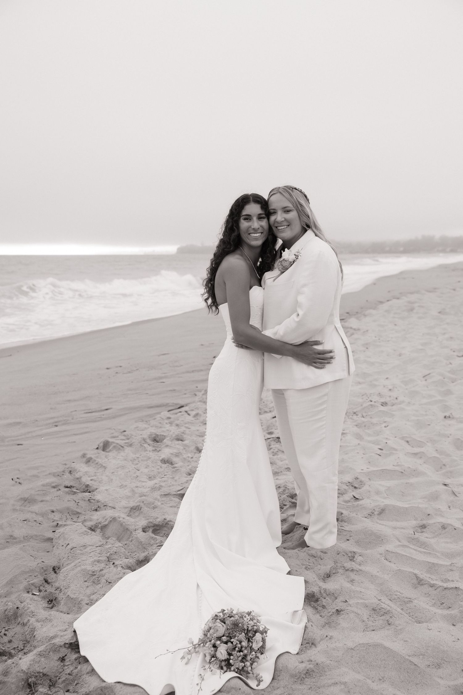 Two brides in white wedding attire embrace on a sandy beach with ocean waves in this black and white photo.
