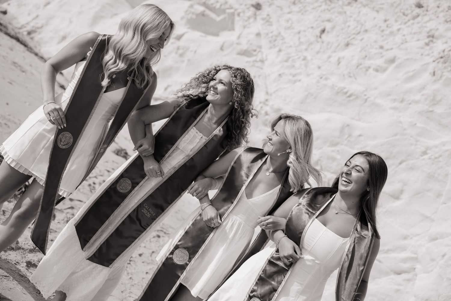Four smiling women wearing graduation stoles pose together on sandy terrain in black and white photo.