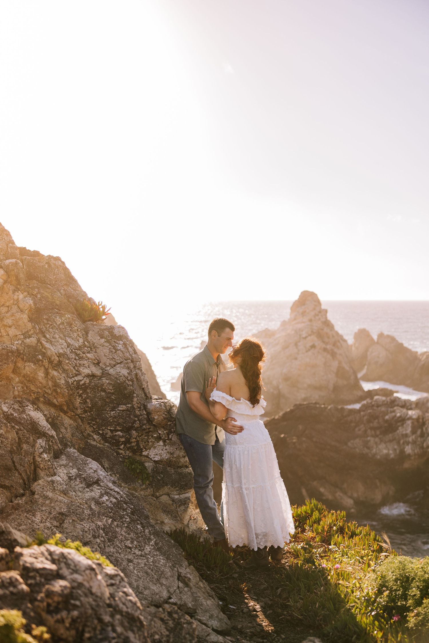 Couple sharing a tender moment on rocky coastal cliffs bathed in warm golden sunset light.
