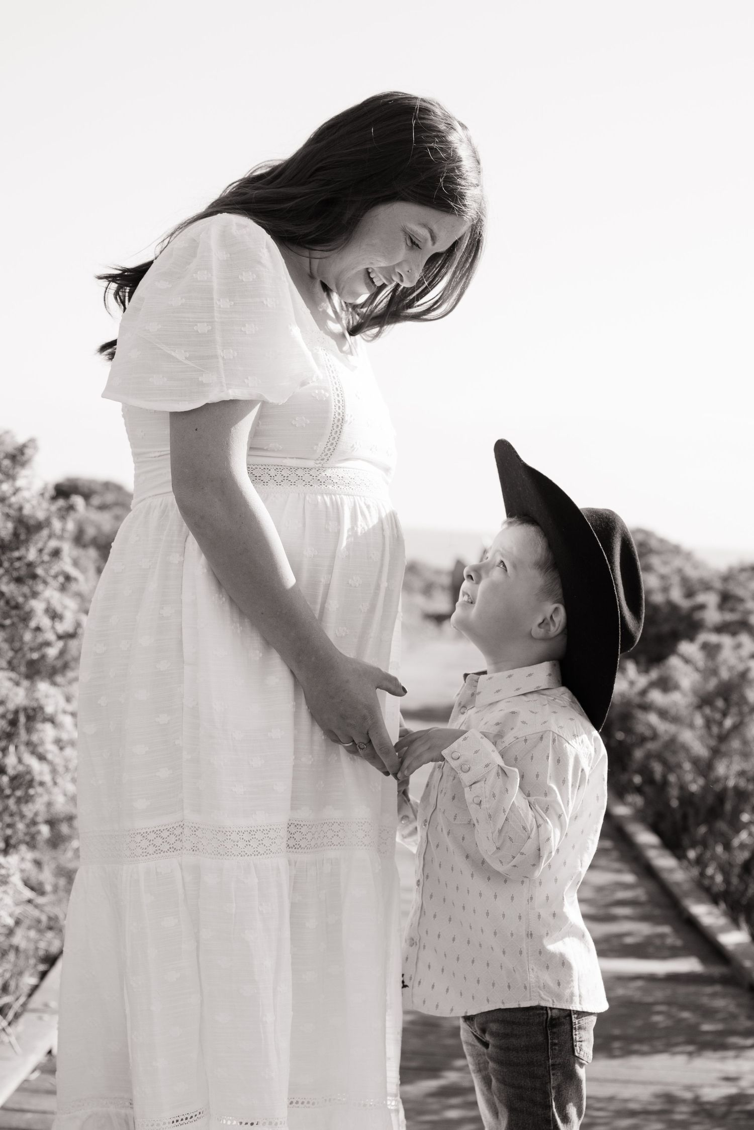 Black and white photo of pregnant woman in white dress smiling at young boy in cowboy hat outdoors.