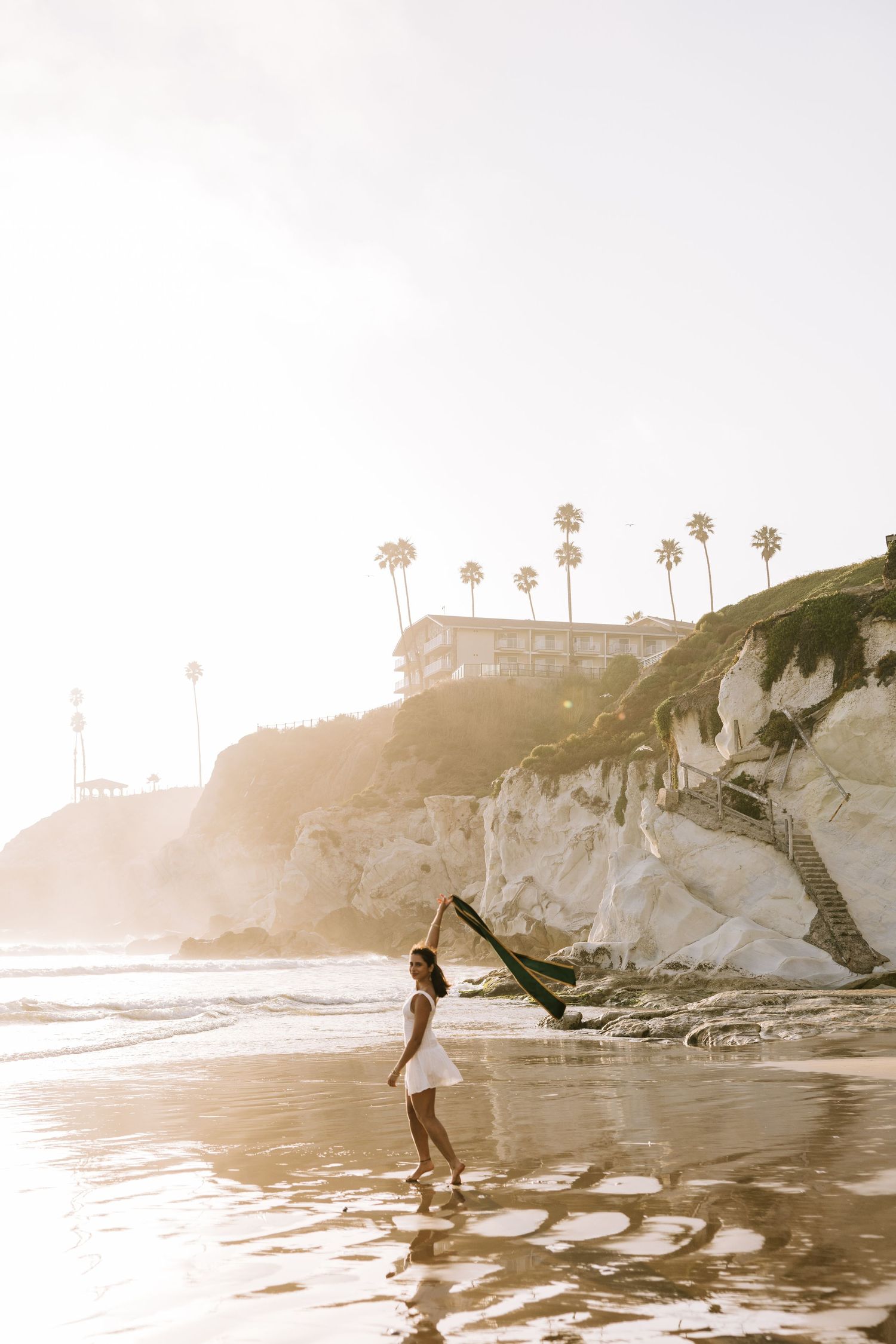 Graduate in white dress waving green stole on sunny beach with palm trees and cliffs.