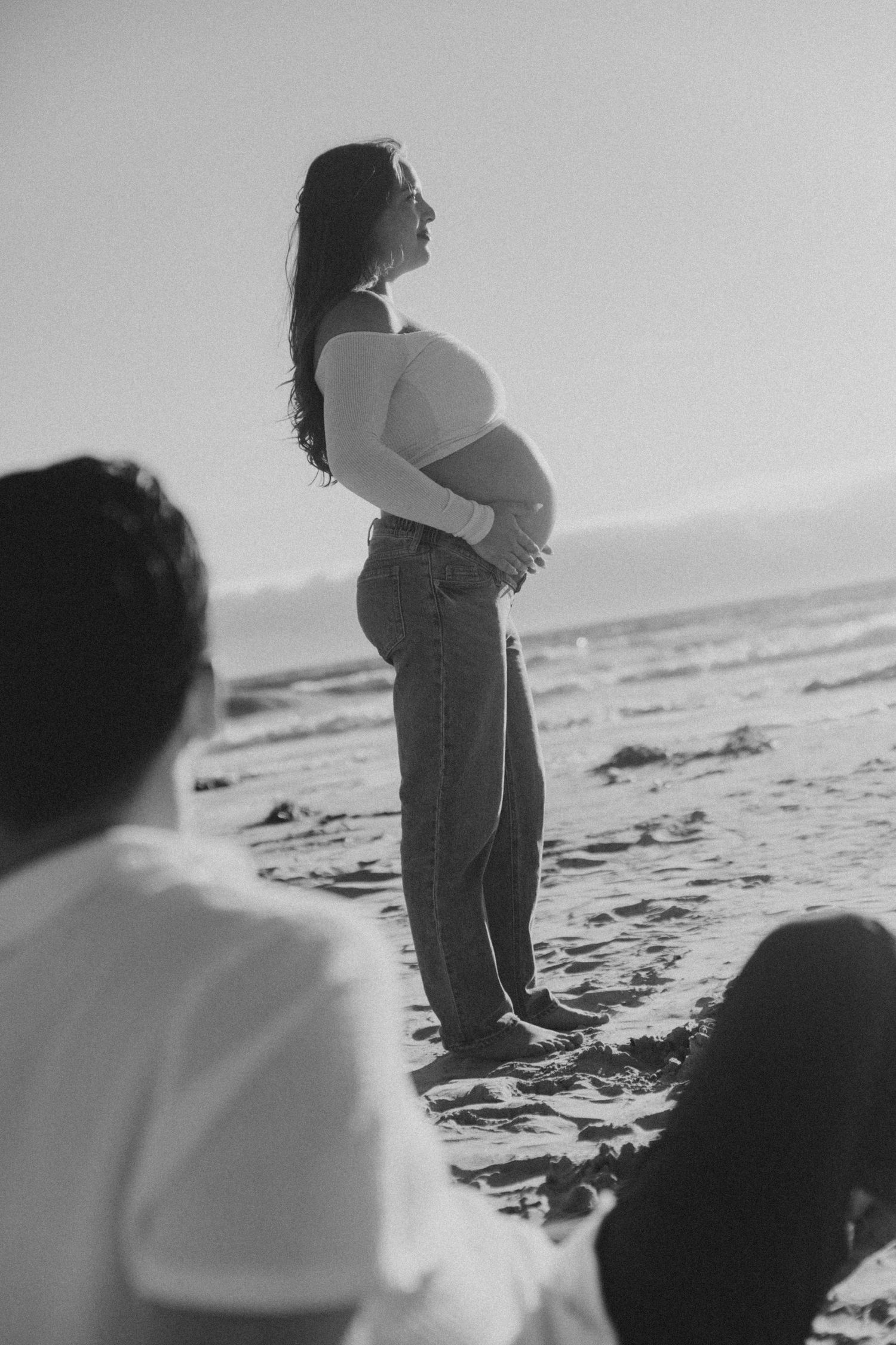 Black and white beach photo of pregnant woman in profile holding baby bump while couple sits nearby.