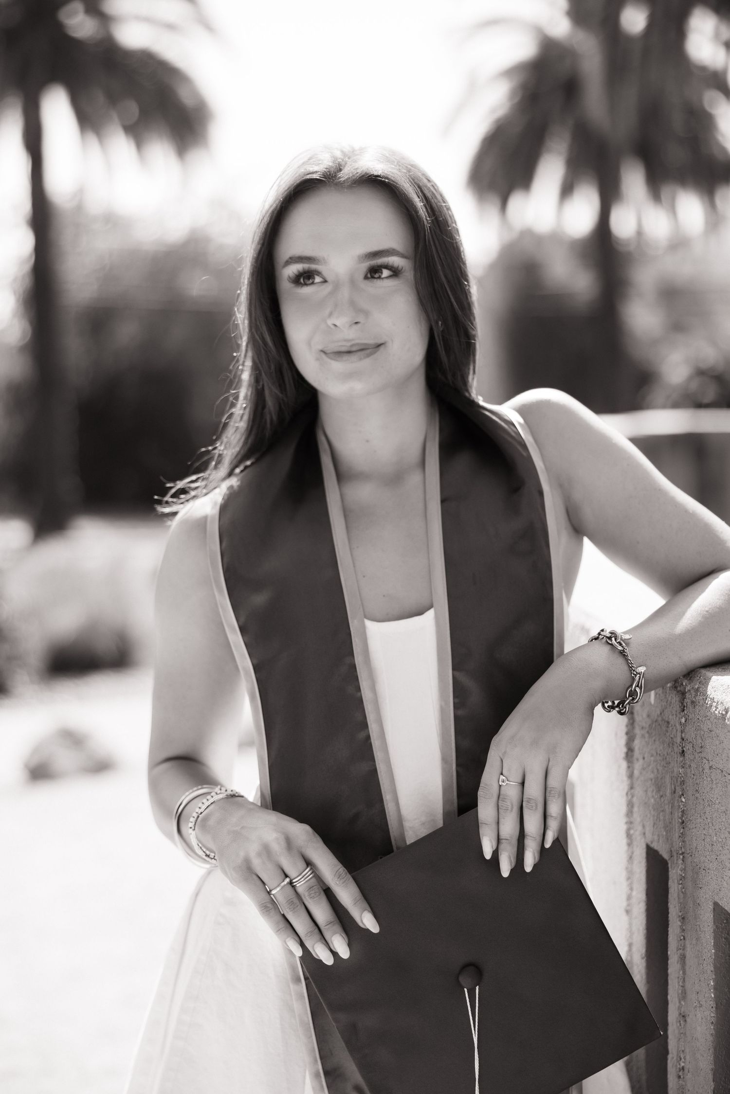 Black and white portrait of brunette woman with graduation cap and stole leaning on railing.