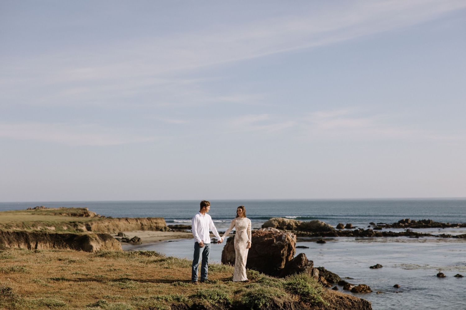 Couple holding hands on rocky coastal shoreline with ocean and blue sky behind them.
