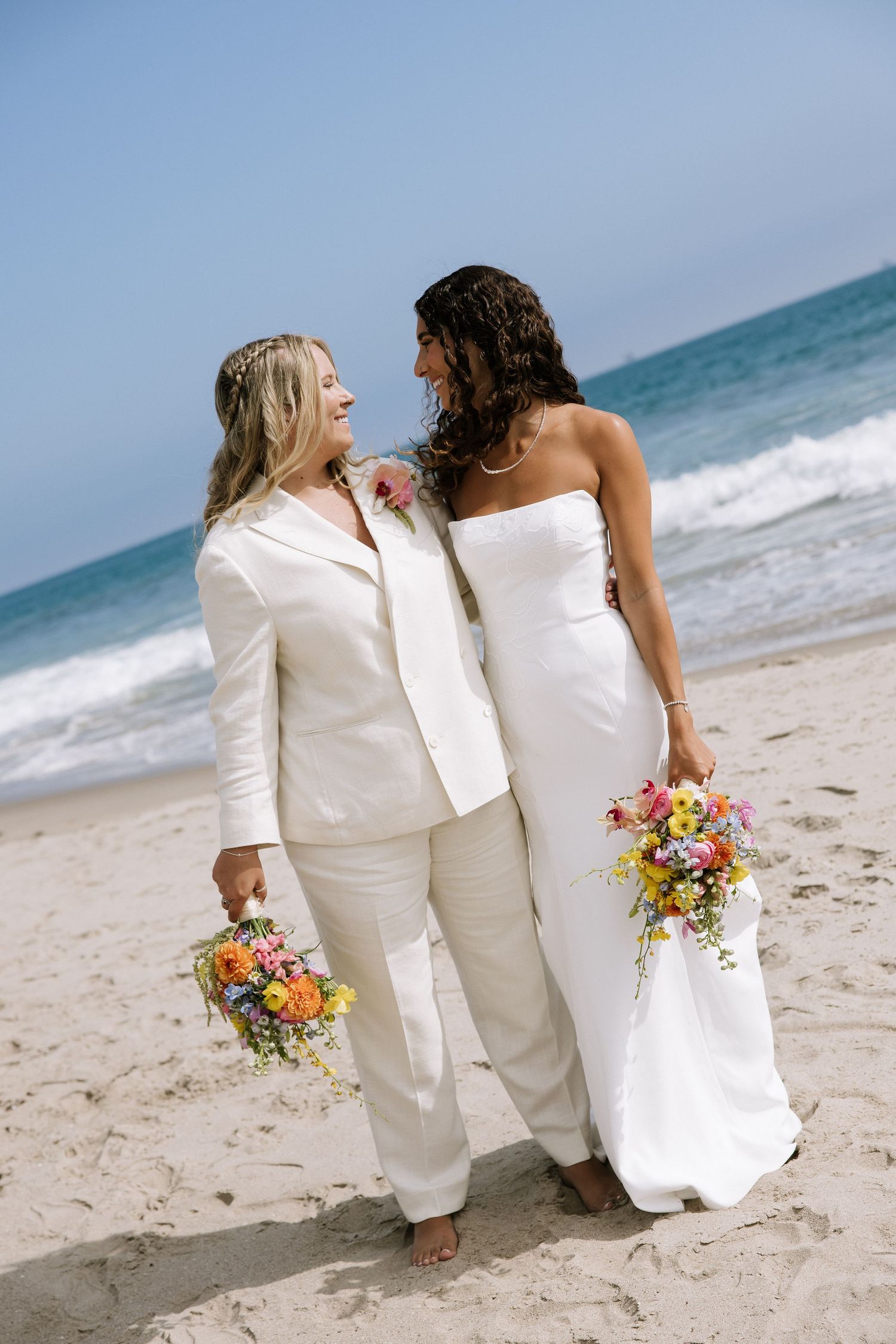 Two brides smile at each other while holding colorful bouquets on a sunny beach.