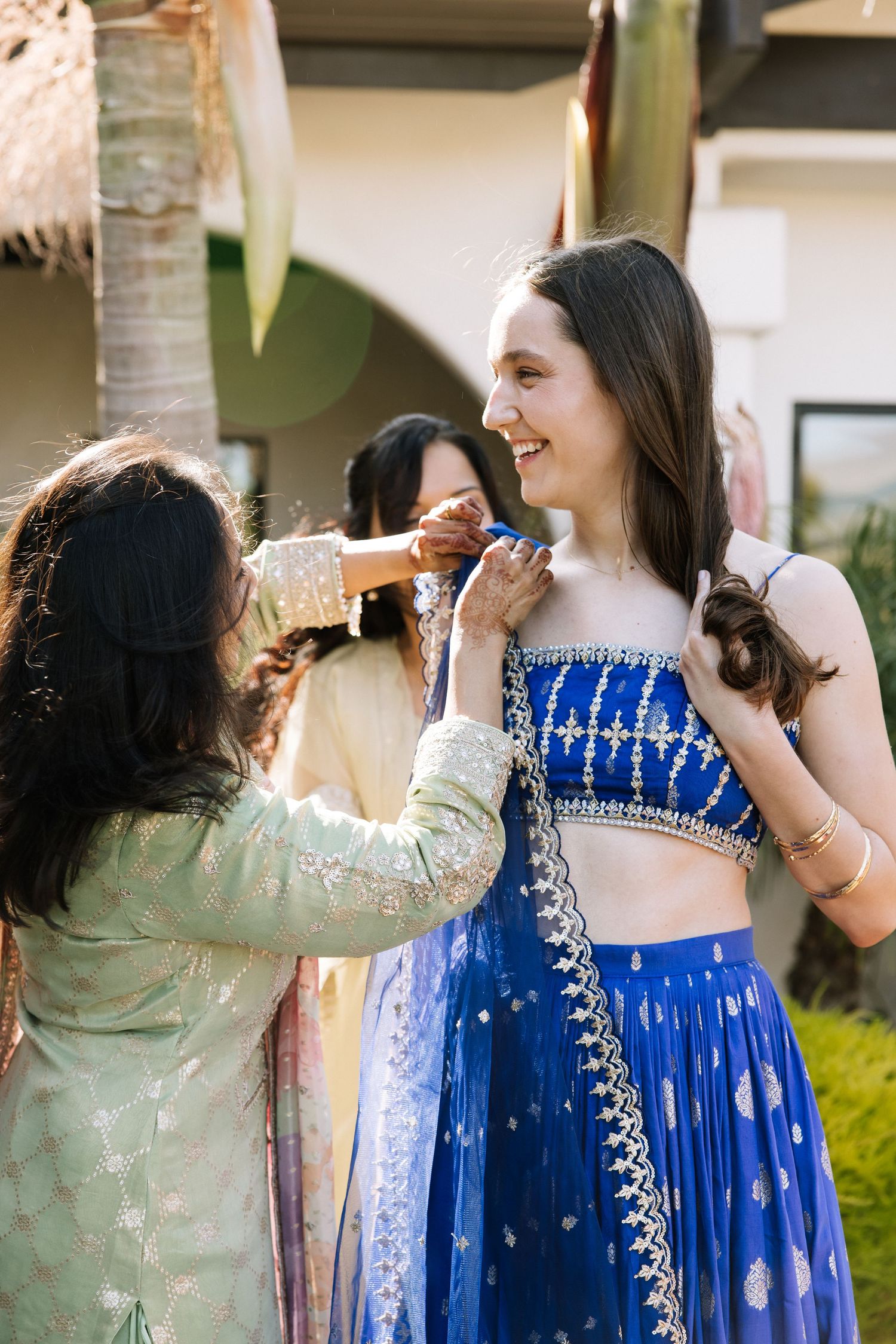 Woman in blue embroidered lehenga smiles as friend adjusts her outfit at colorful Indian wedding celebration.