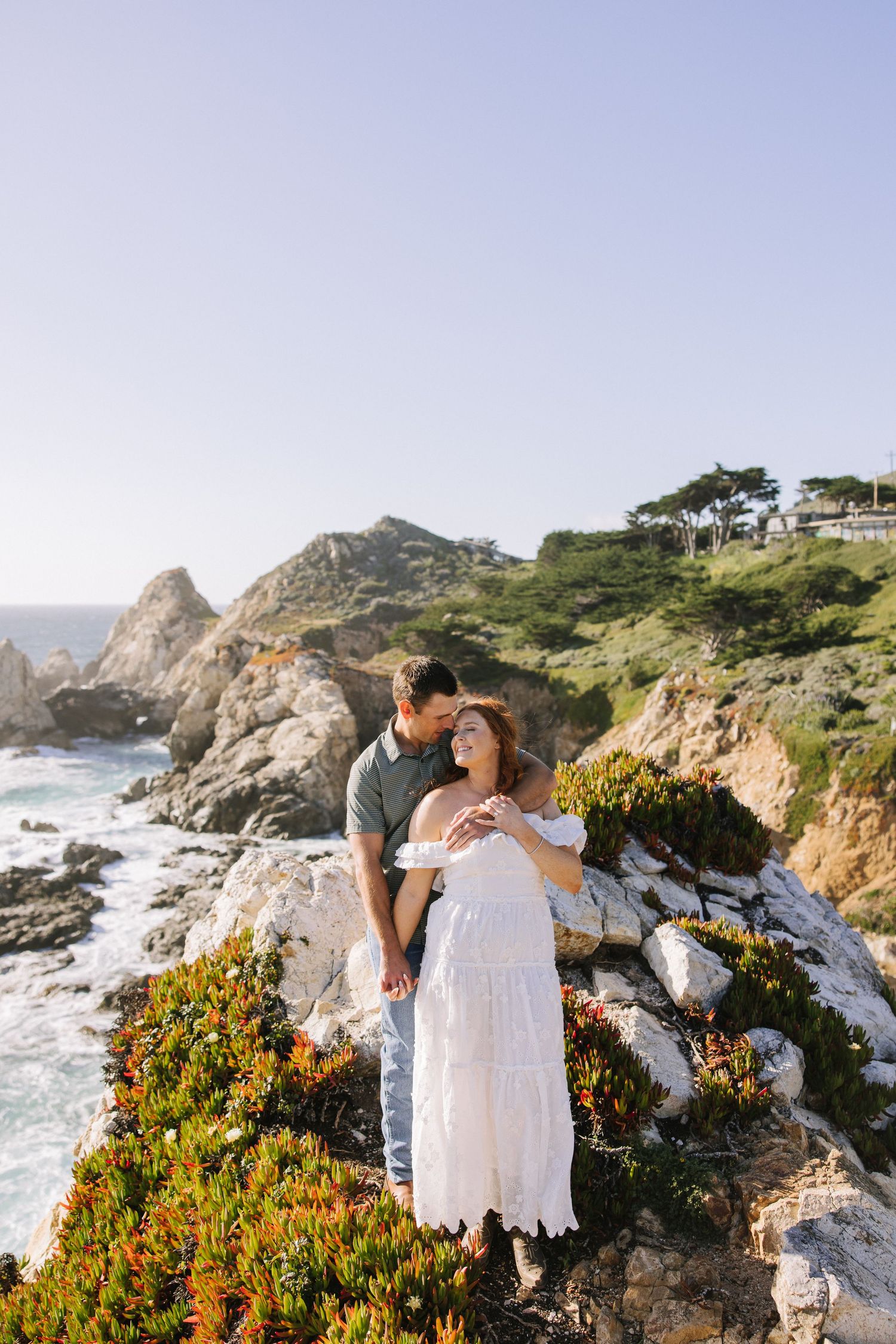 Romantic couple embracing on scenic coastal cliffs with ocean views and colorful wildflowers.