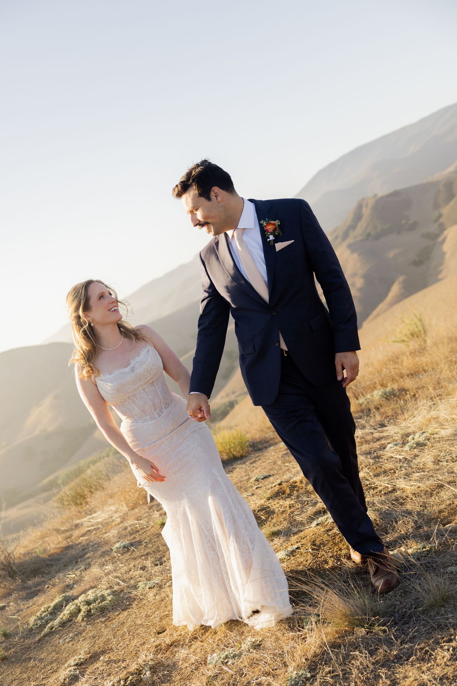 Bride and groom walk hand in hand through golden hillside with mountain backdrop on their wedding day.