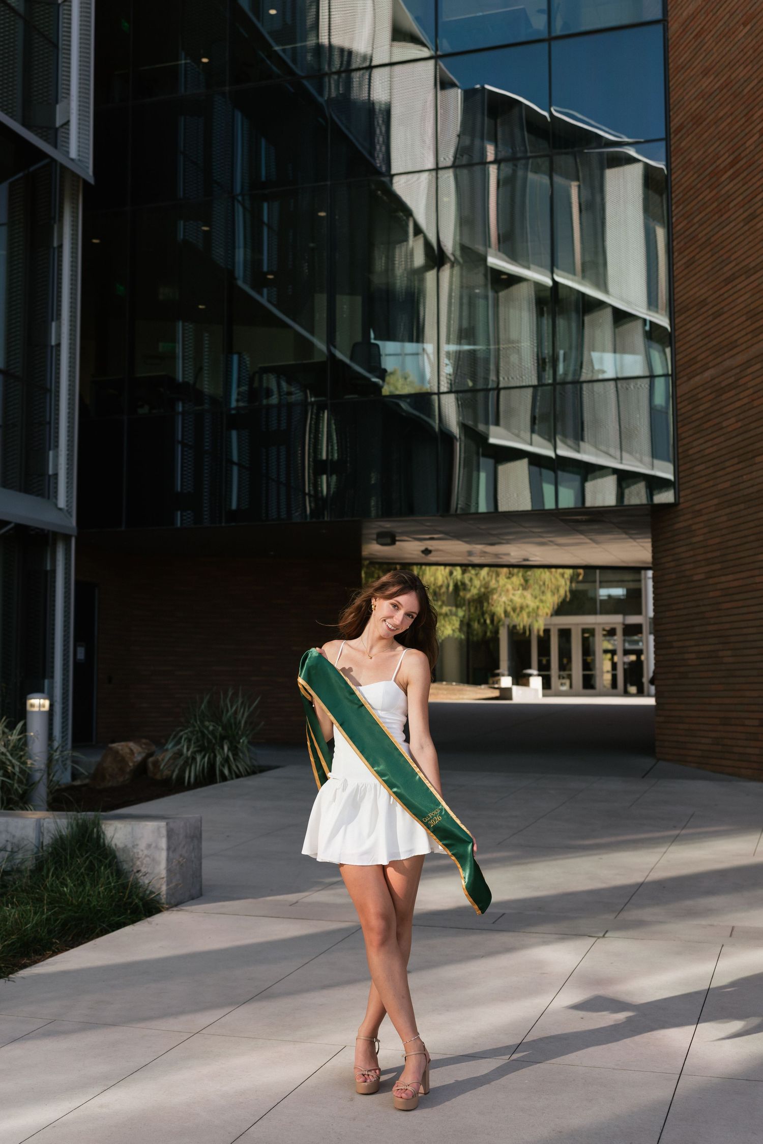 Young woman in white dress wearing green graduation sash posing outside modern university building.