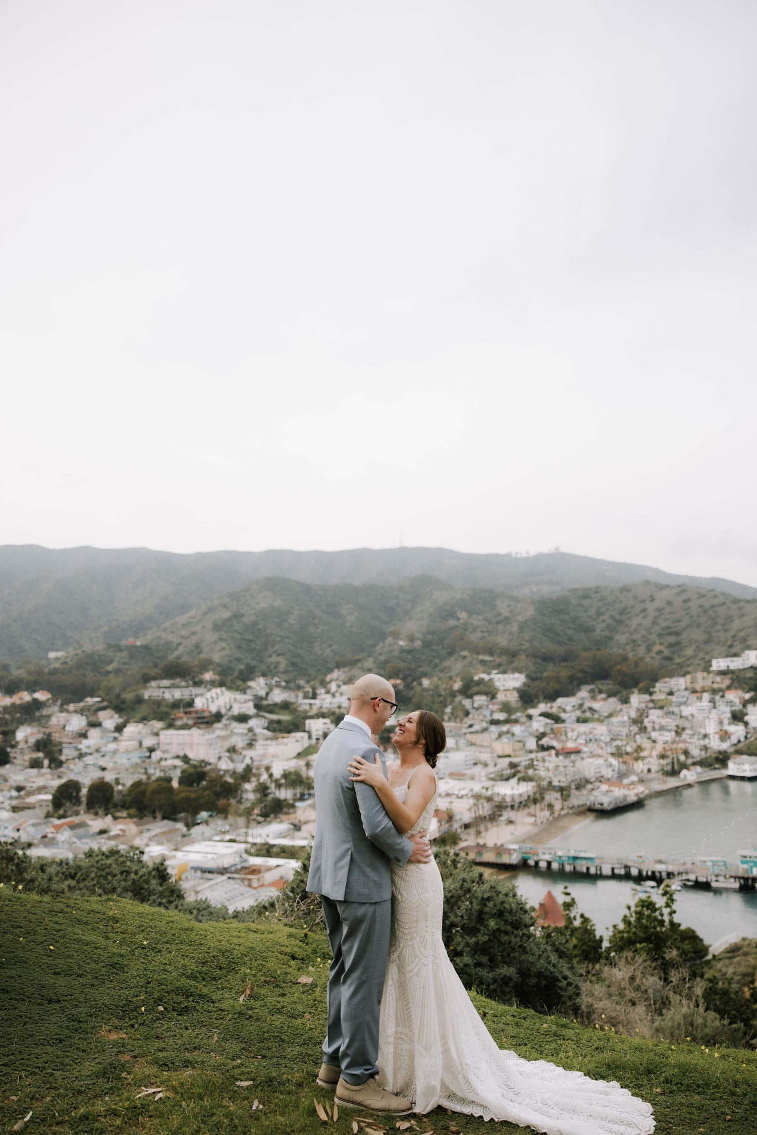 Bride and groom embrace on a hilltop overlooking a coastal town with mountains in the background on their wedding day.