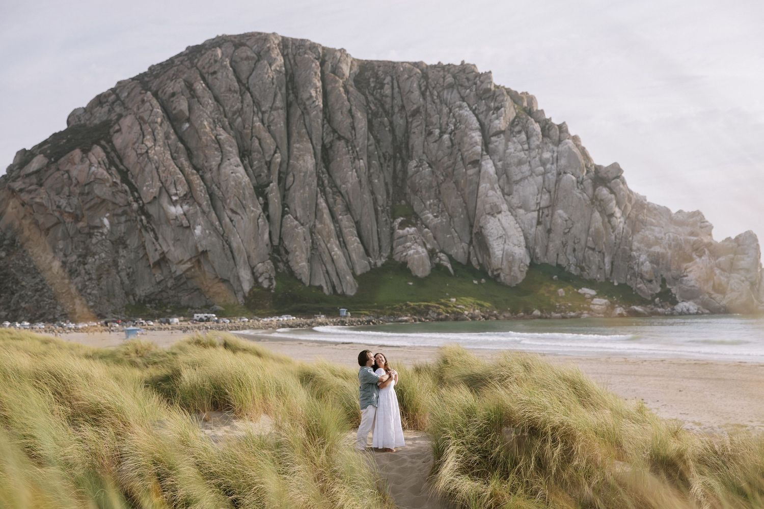 Couple embracing in wedding attire among coastal dunes with dramatic Morro Rock in the background.