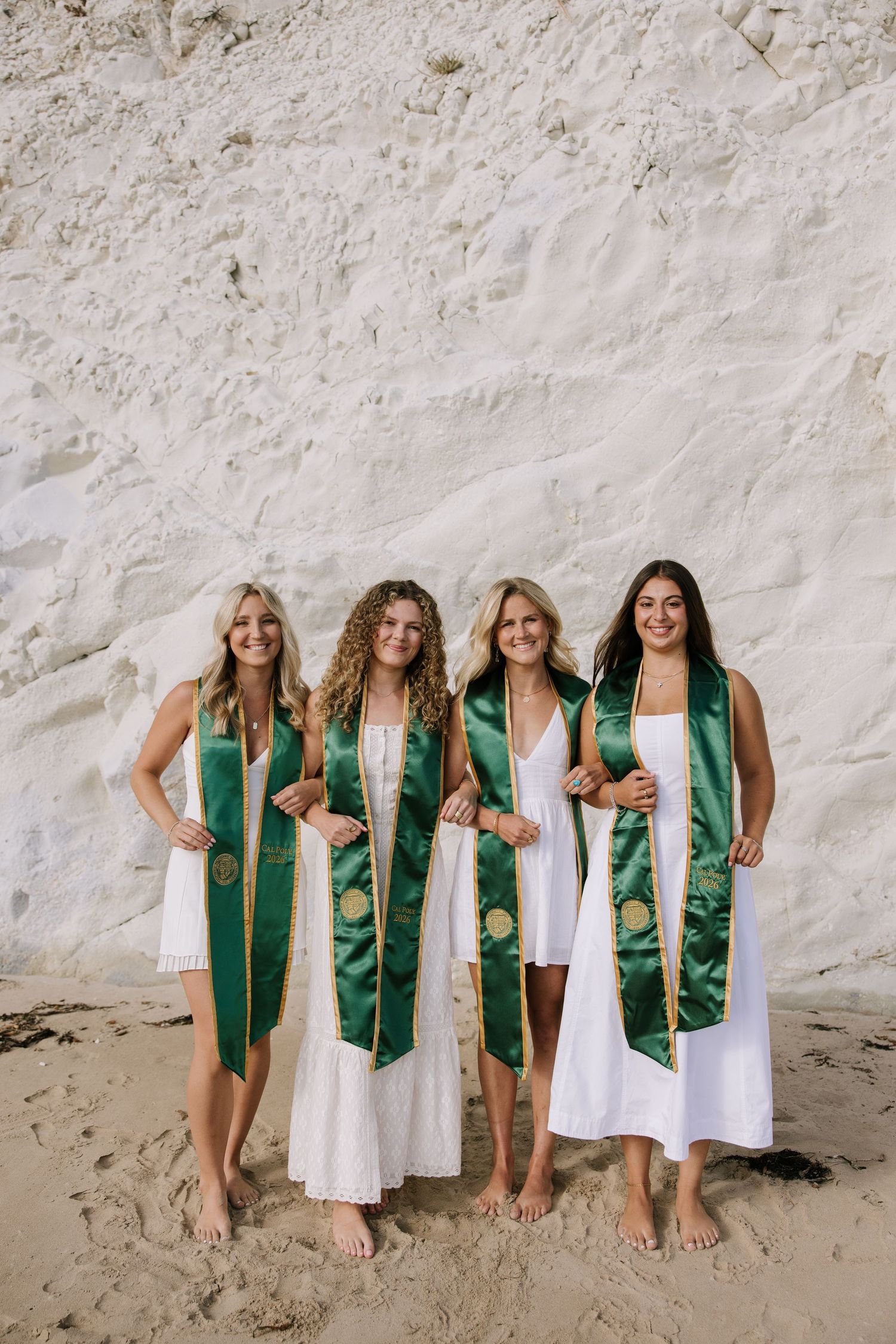 Four female graduates wearing green sashes pose barefoot on sandy beach in white dresses.