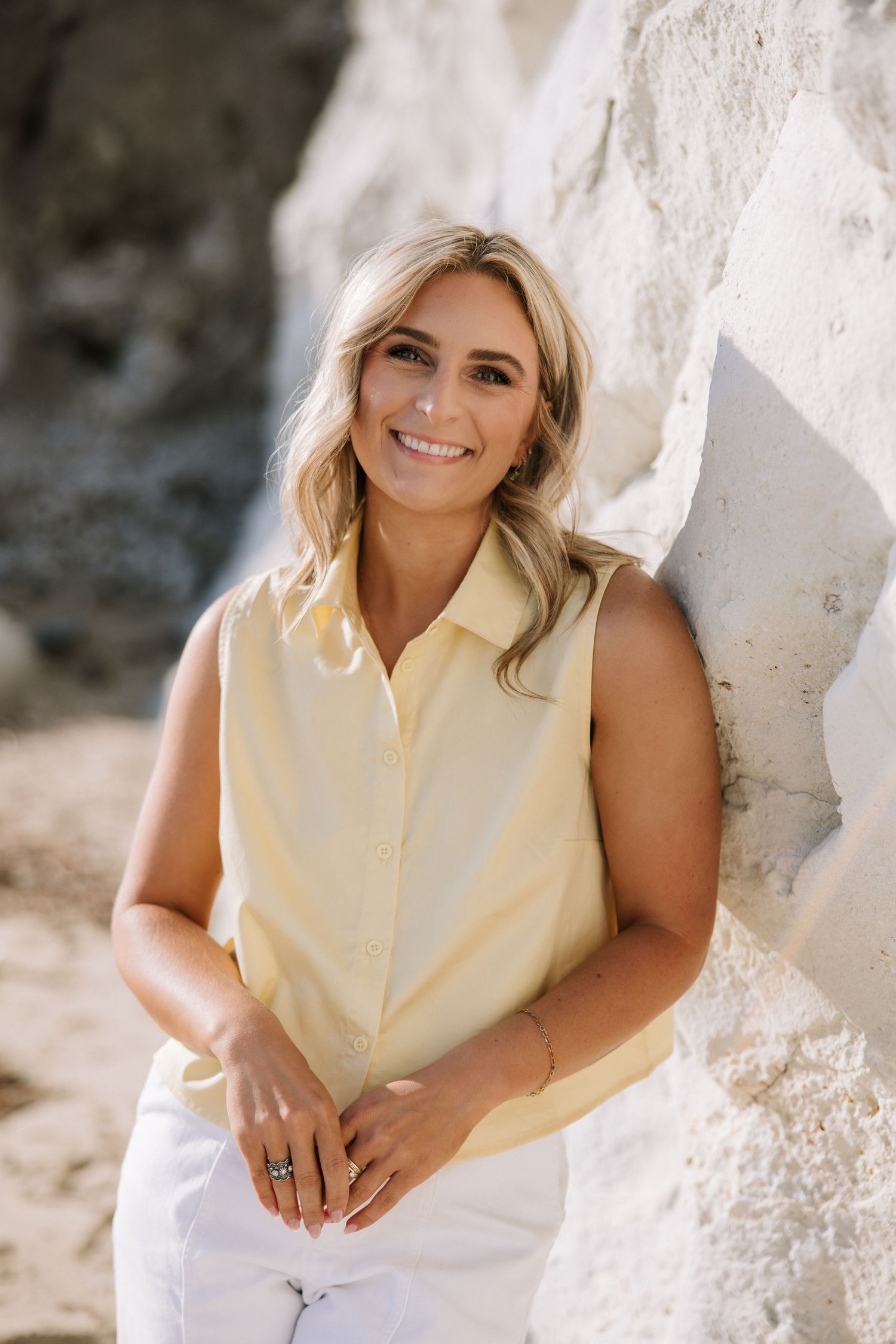 Blonde woman in yellow sleeveless shirt leaning against white rock wall smiling for portrait photos.