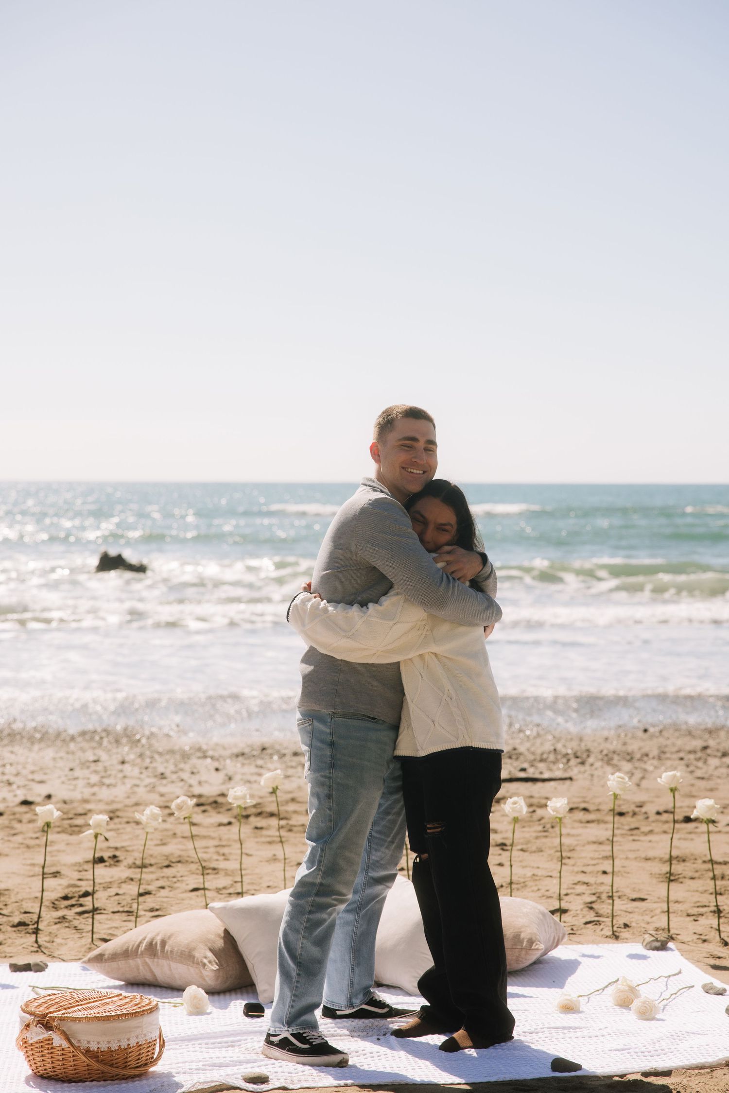 Newly engaged couple hugging on a beach blanket surrounded by white roses and a picnic basket.