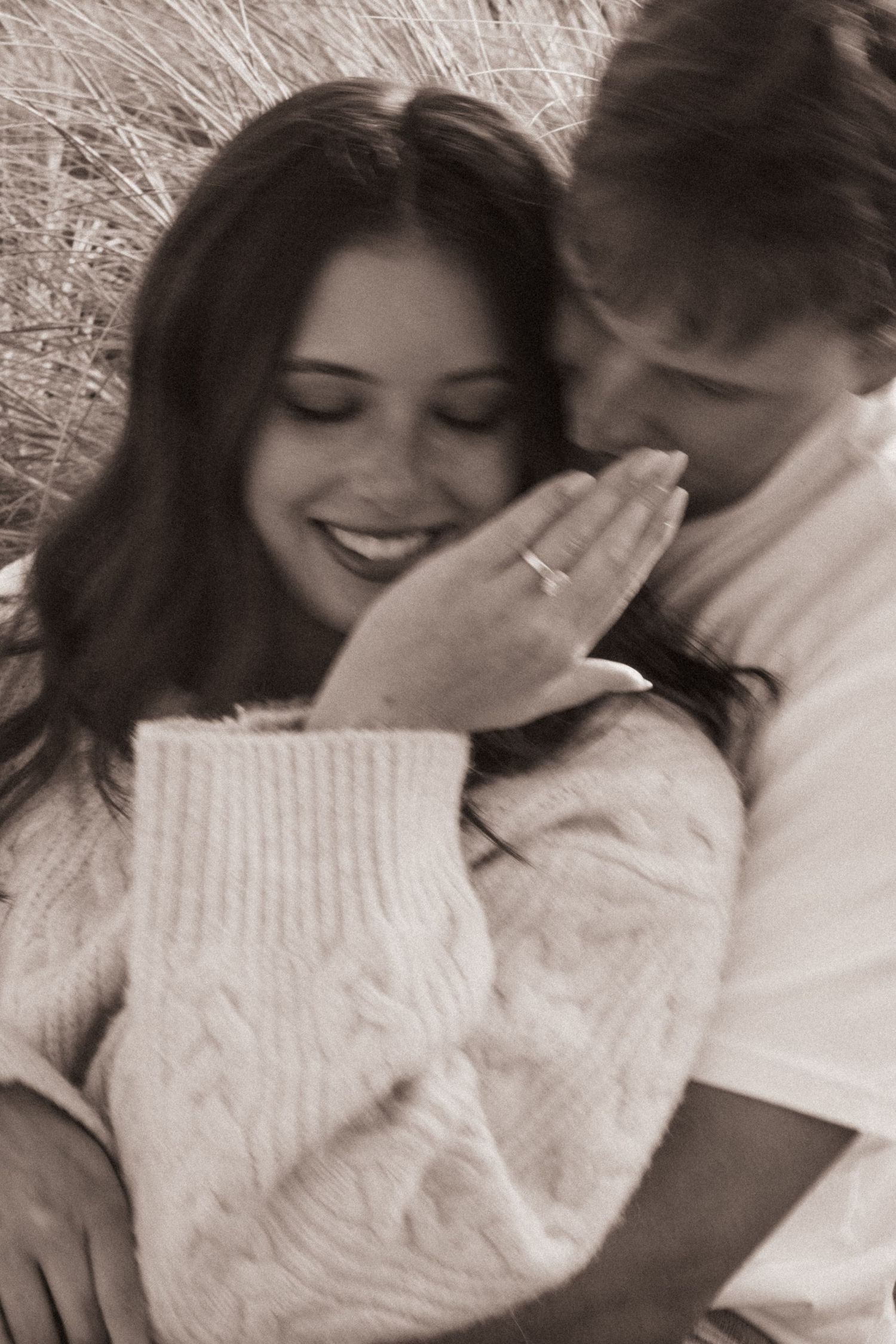 Sepia toned photo of smiling woman with engagement ring snuggled close to partner in field.