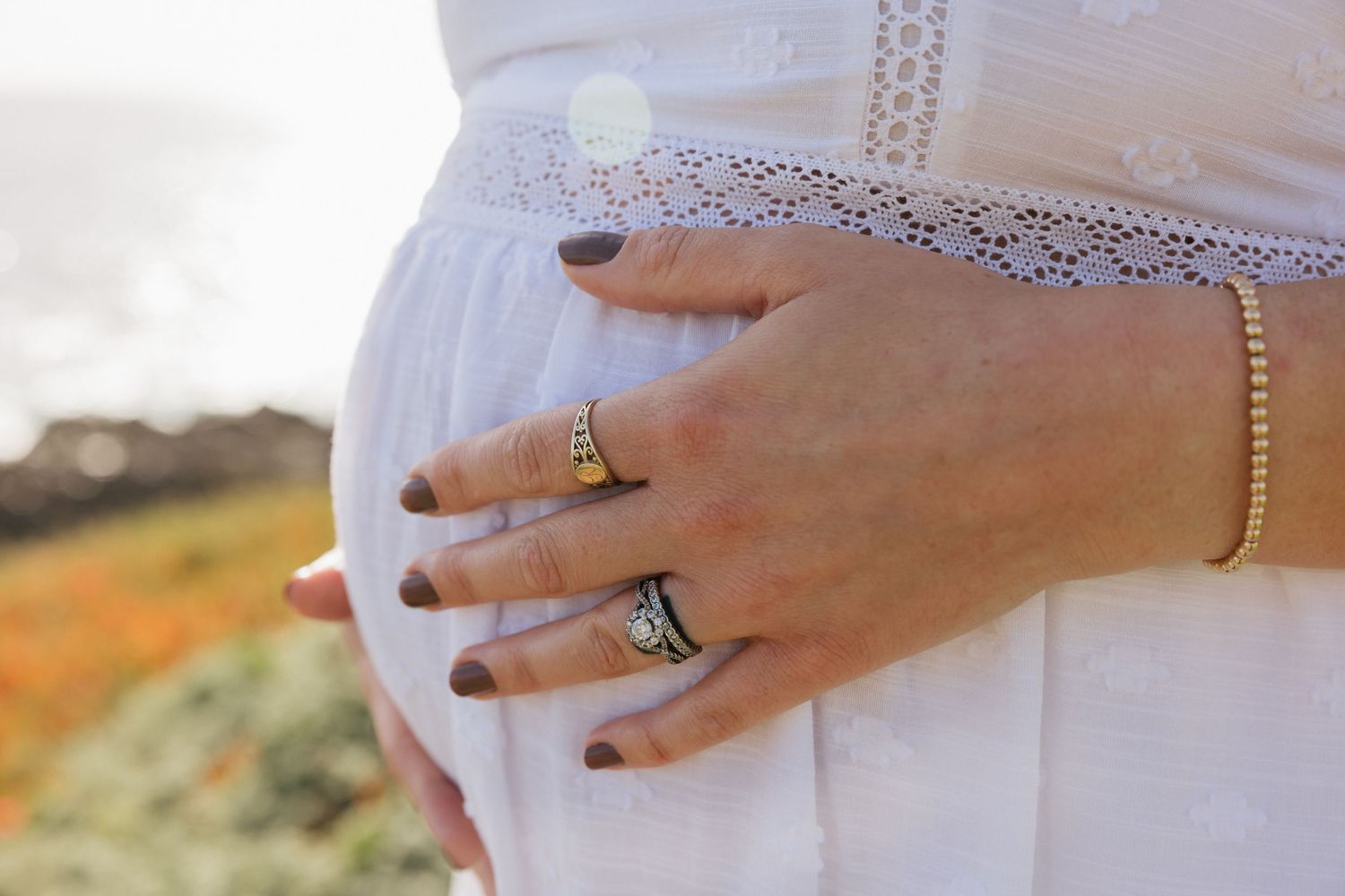 Close-up of pregnant belly in white lace dress with rings and gold bracelet on hands.