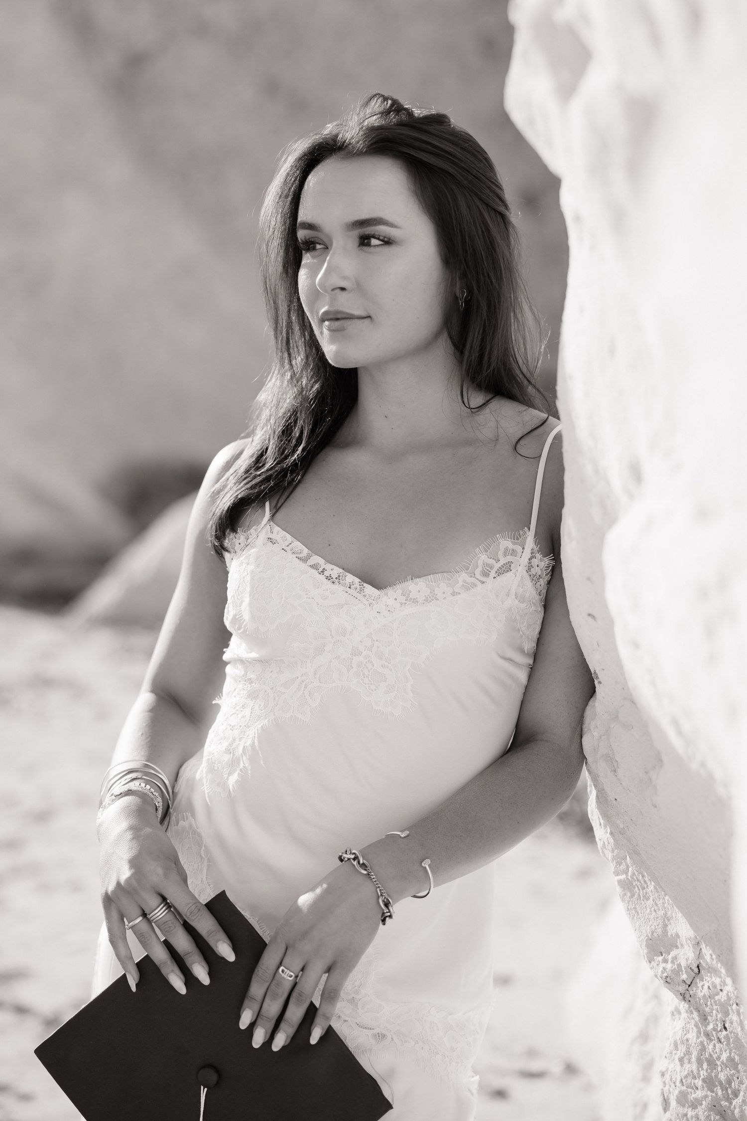 Black and white portrait of brunette woman in white dress holding graduation cap by rock.