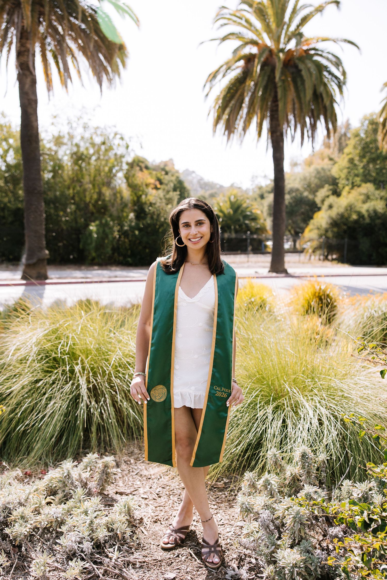 Smiling graduate in white dress and green stole standing among plants with palm trees behind.