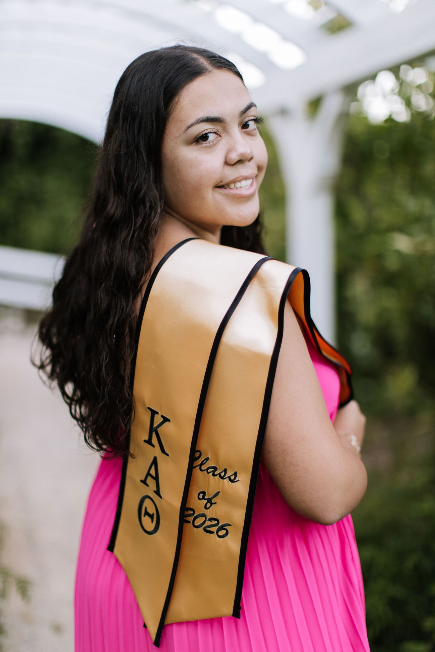 Smiling woman in pink dress wearing gold Kappa Alpha Theta Class of 2026 graduation sash outdoors.
