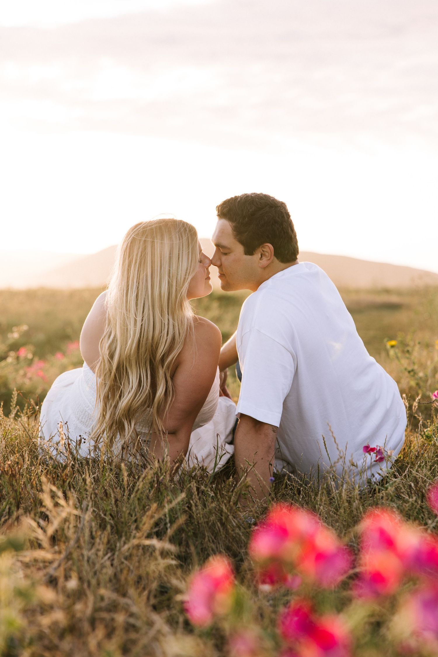 Couple shares a kiss while sitting in wildflower meadow during golden hour sunset.