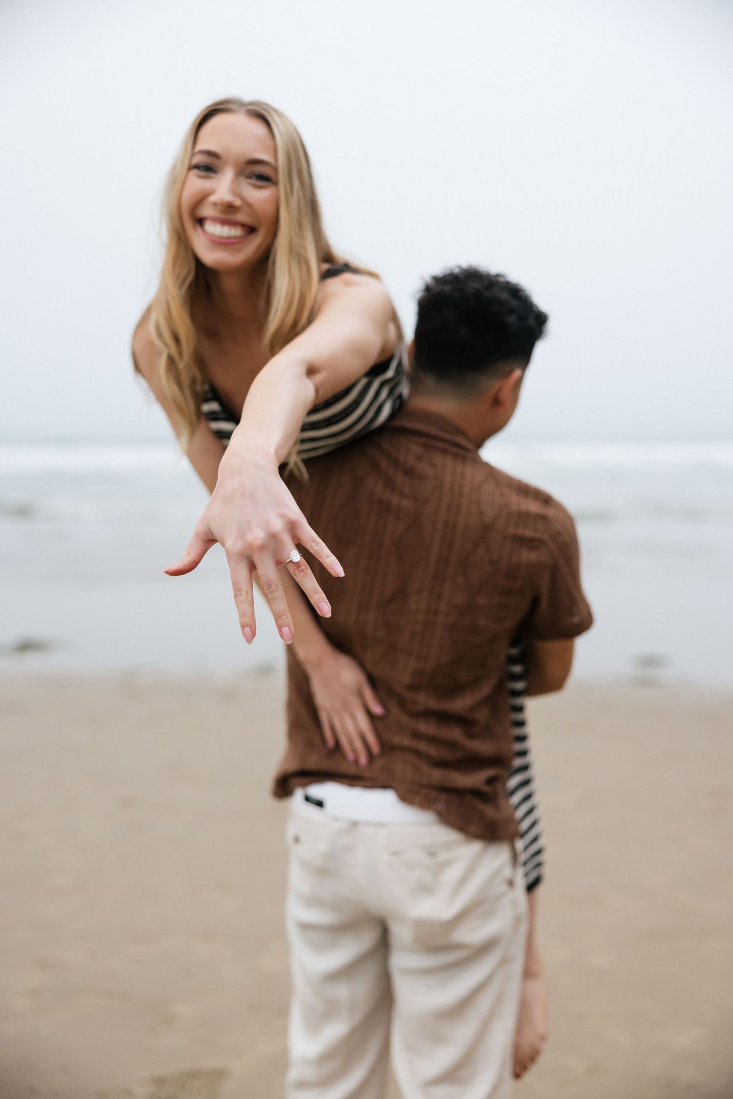 Joyful woman showing off her engagement ring while being lifted by her partner on a sandy beach.