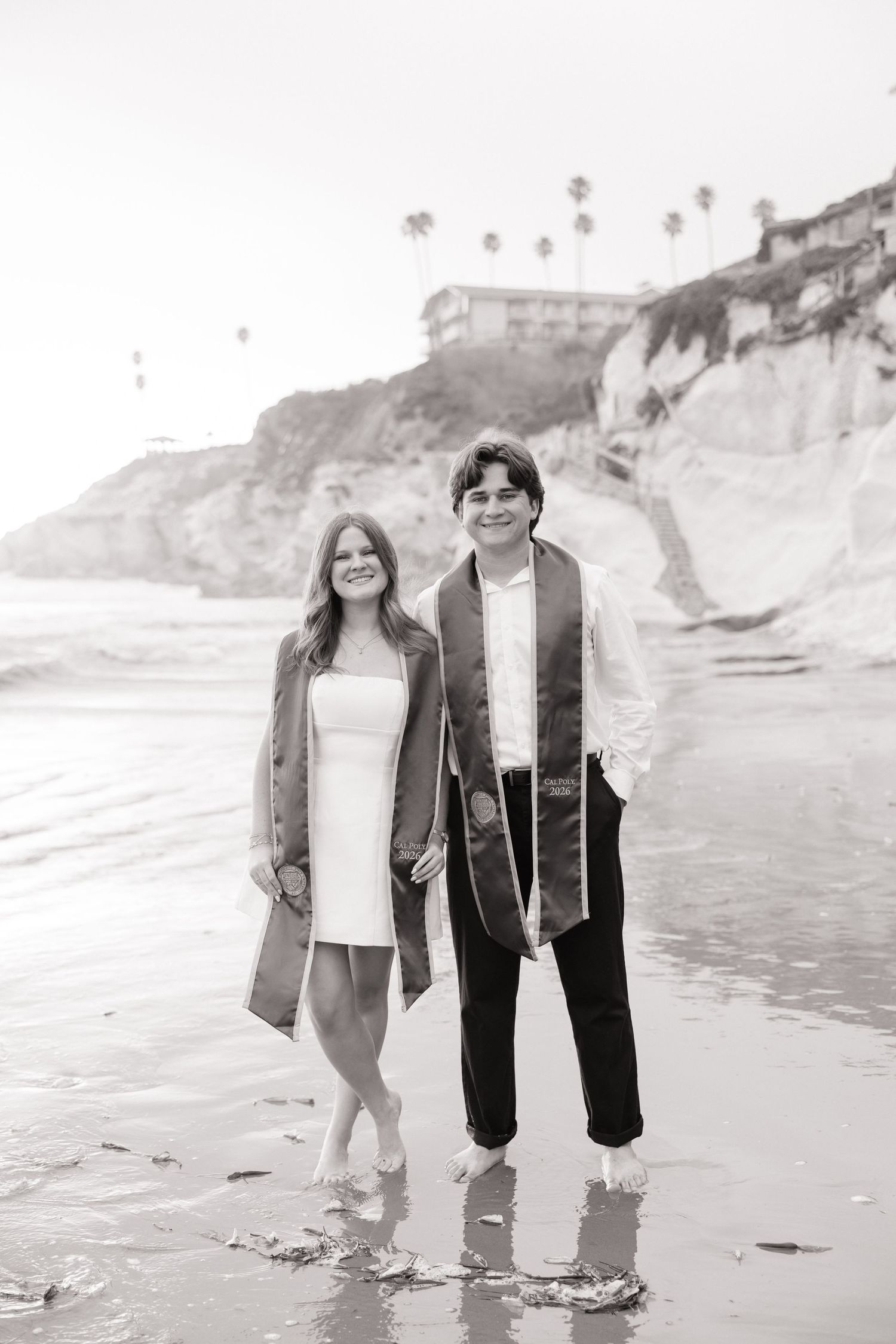 Black and white photo of a smiling couple in graduation attire walking barefoot on a beach with cliffs behind them.