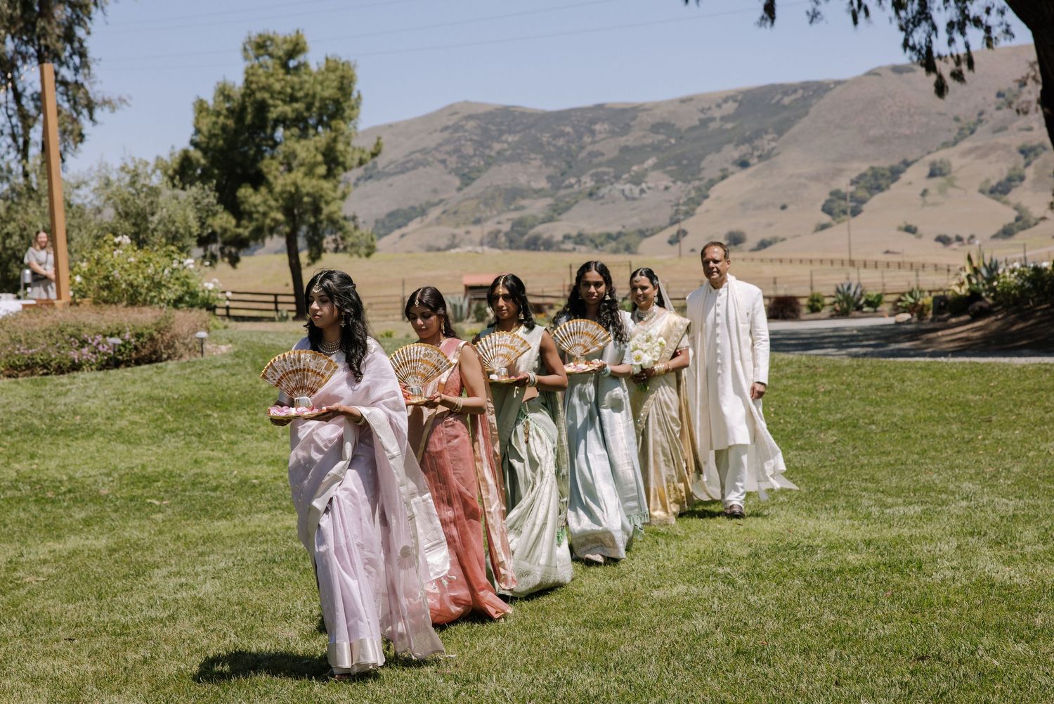Bridesmaids in pastel sarees walking across green lawn with scenic mountain backdrop.