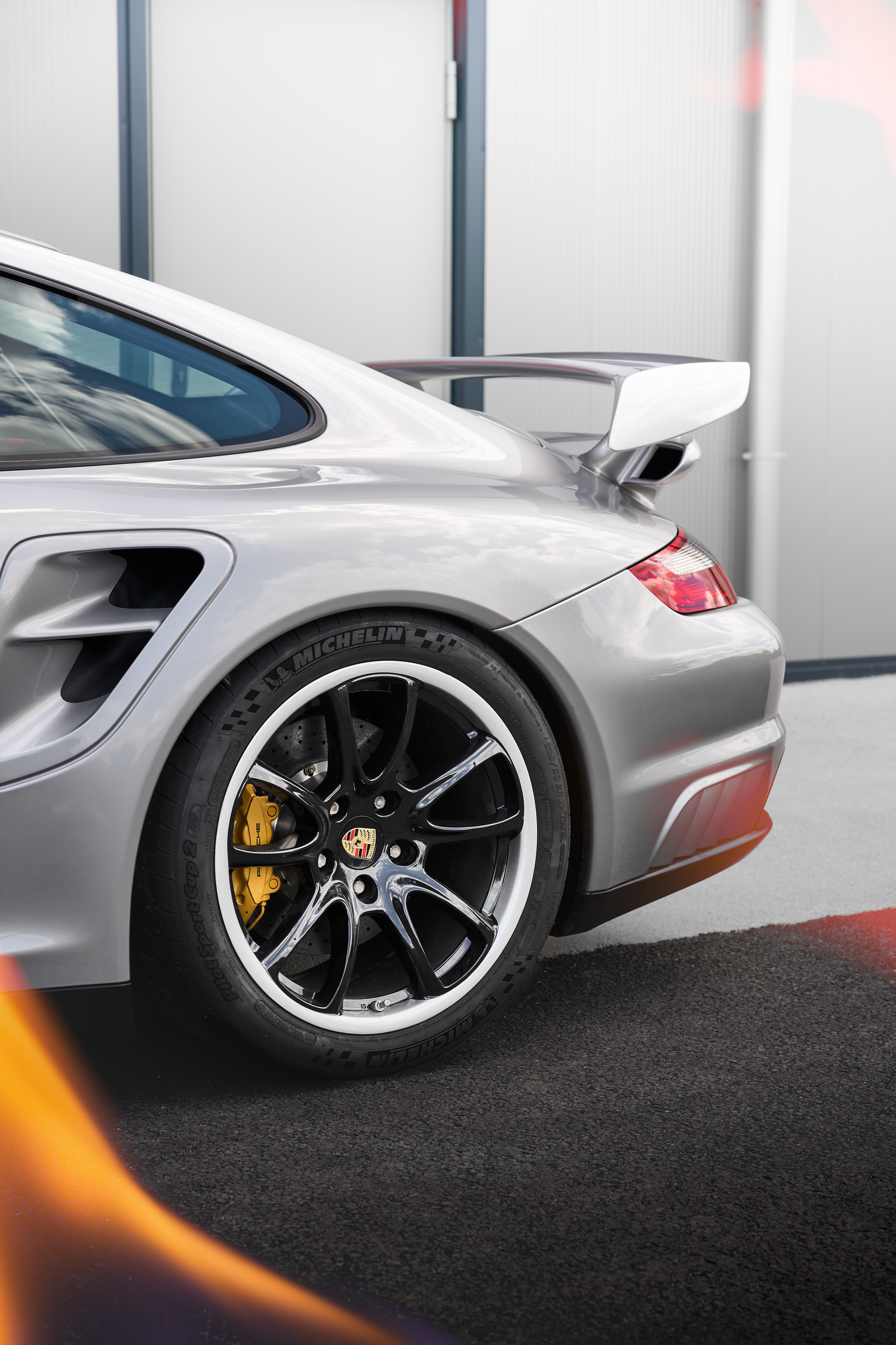 Silver Porsche 911 GT2 RS rear view with yellow brake calipers, black alloy wheels, and rear spoiler in a garage.