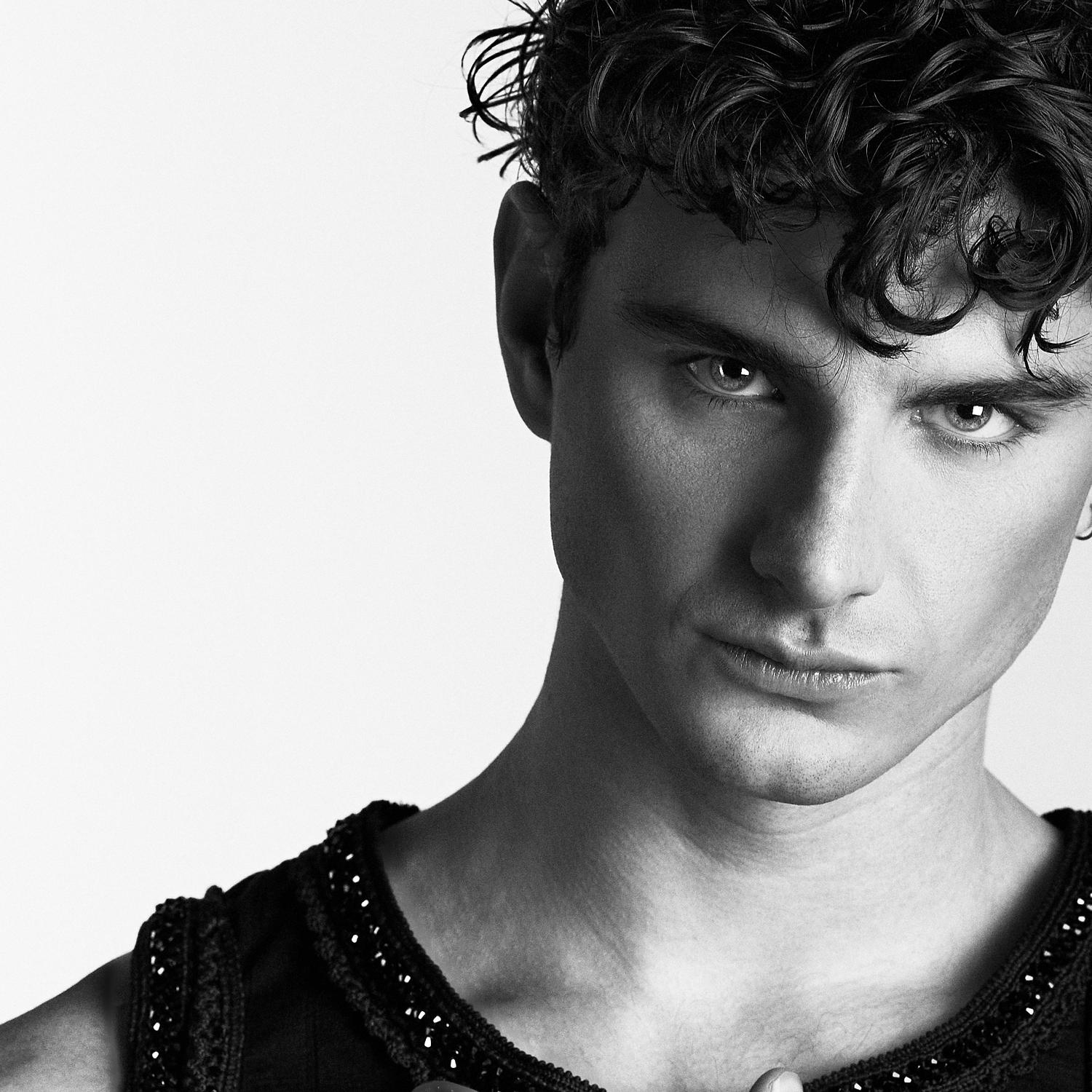 Black and white portrait of a young man with curly hair wearing a black sleeveless top, intense gaze.