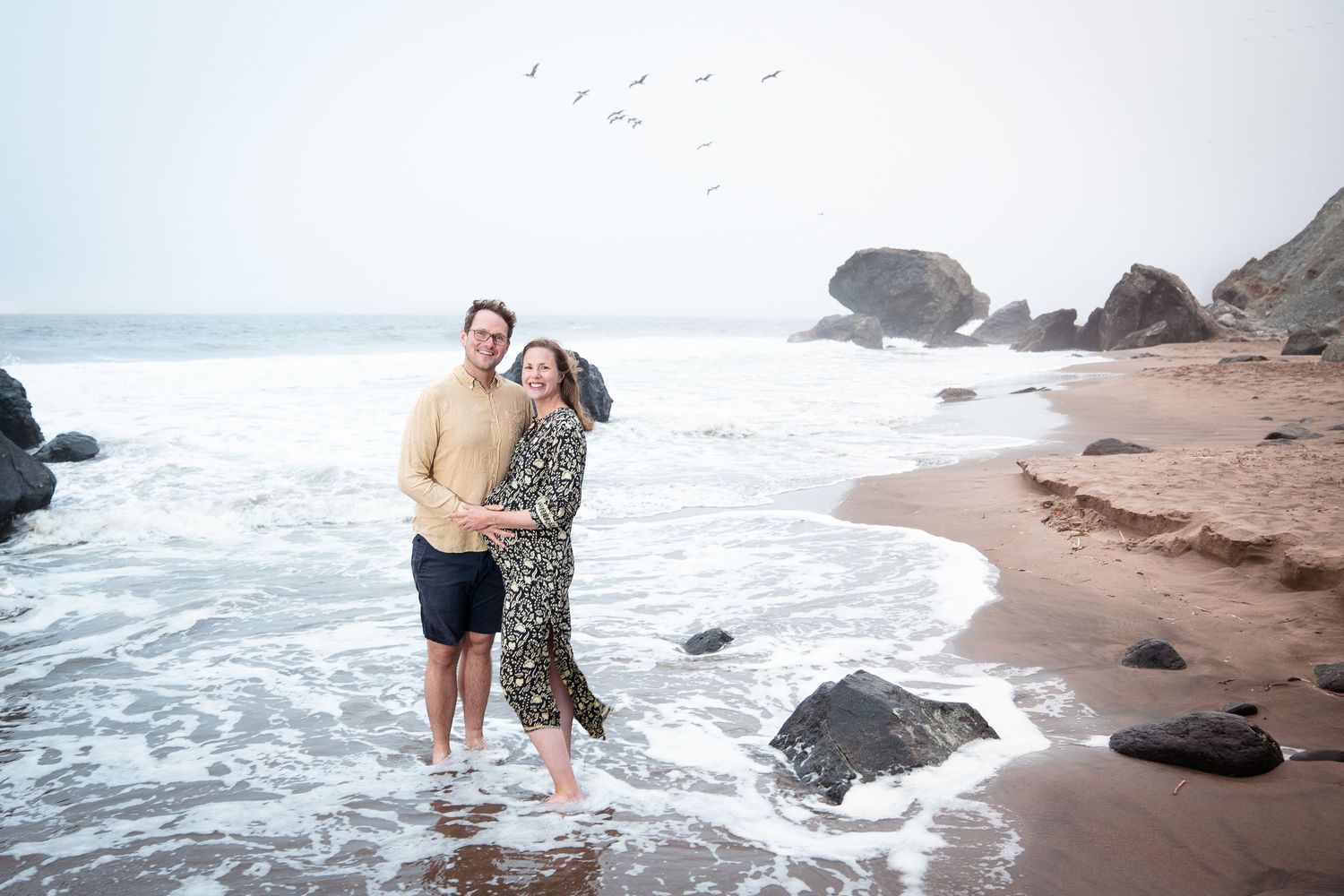 A couple stands together on a rocky beach with crashing waves and seabirds flying overhead.
