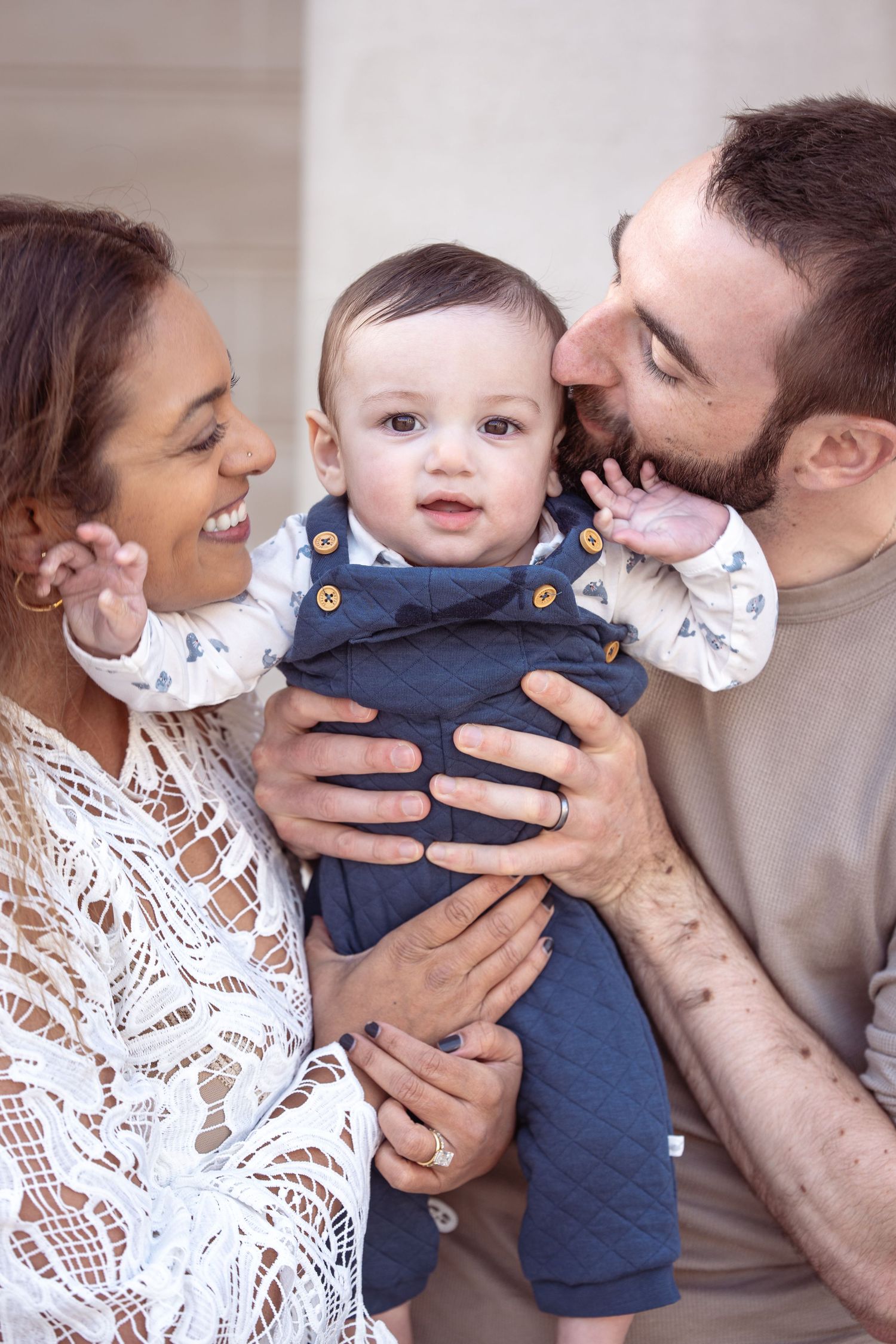 A loving family moment showing parents holding and kissing their baby wearing a navy blue outfit.