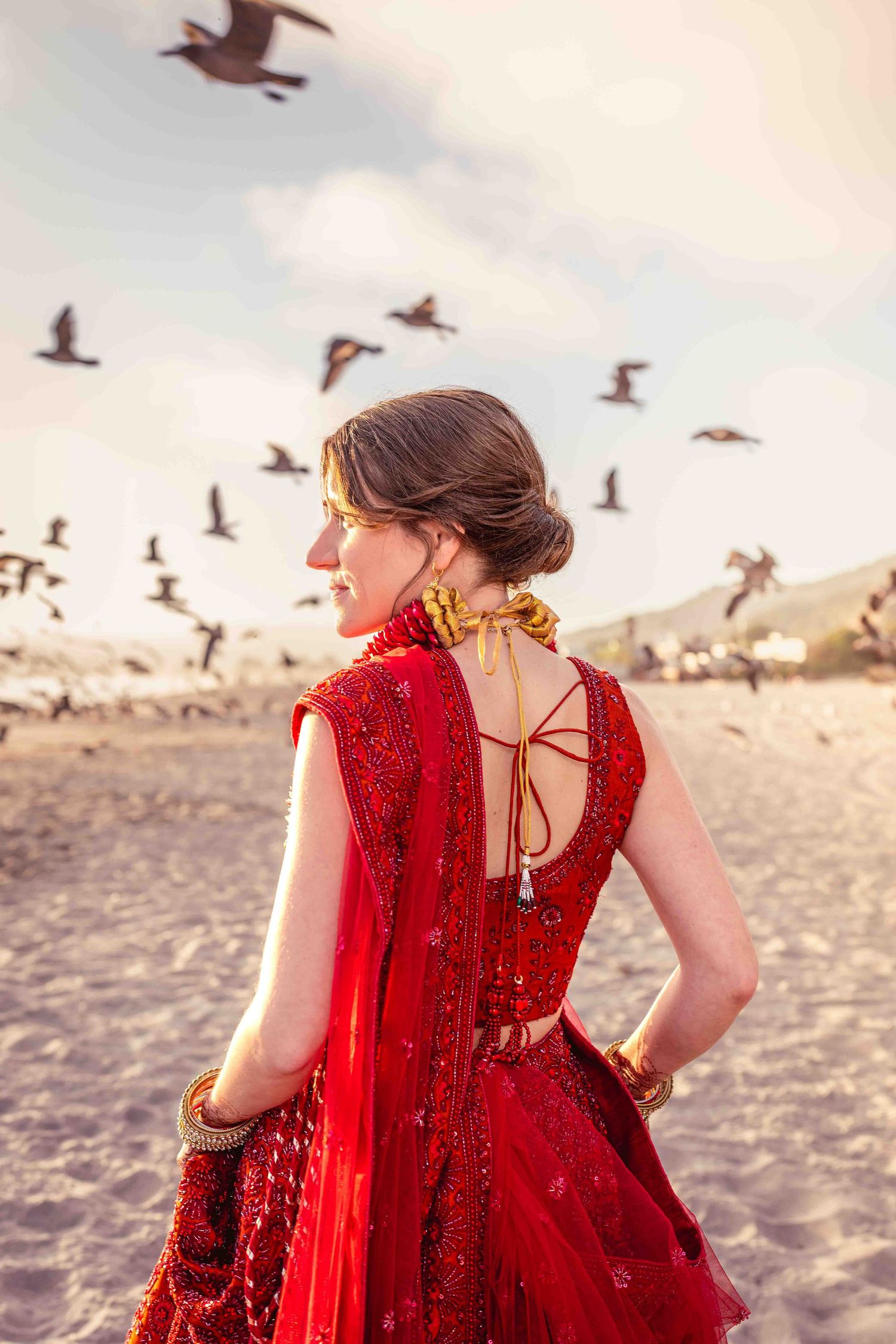 A person in red attire photographed from behind as seabirds fly overhead on a beach.