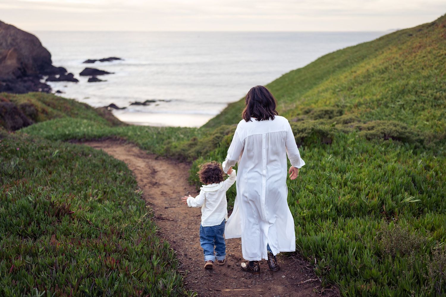 Parent and child walking hand in hand down coastal trail overlooking ocean.