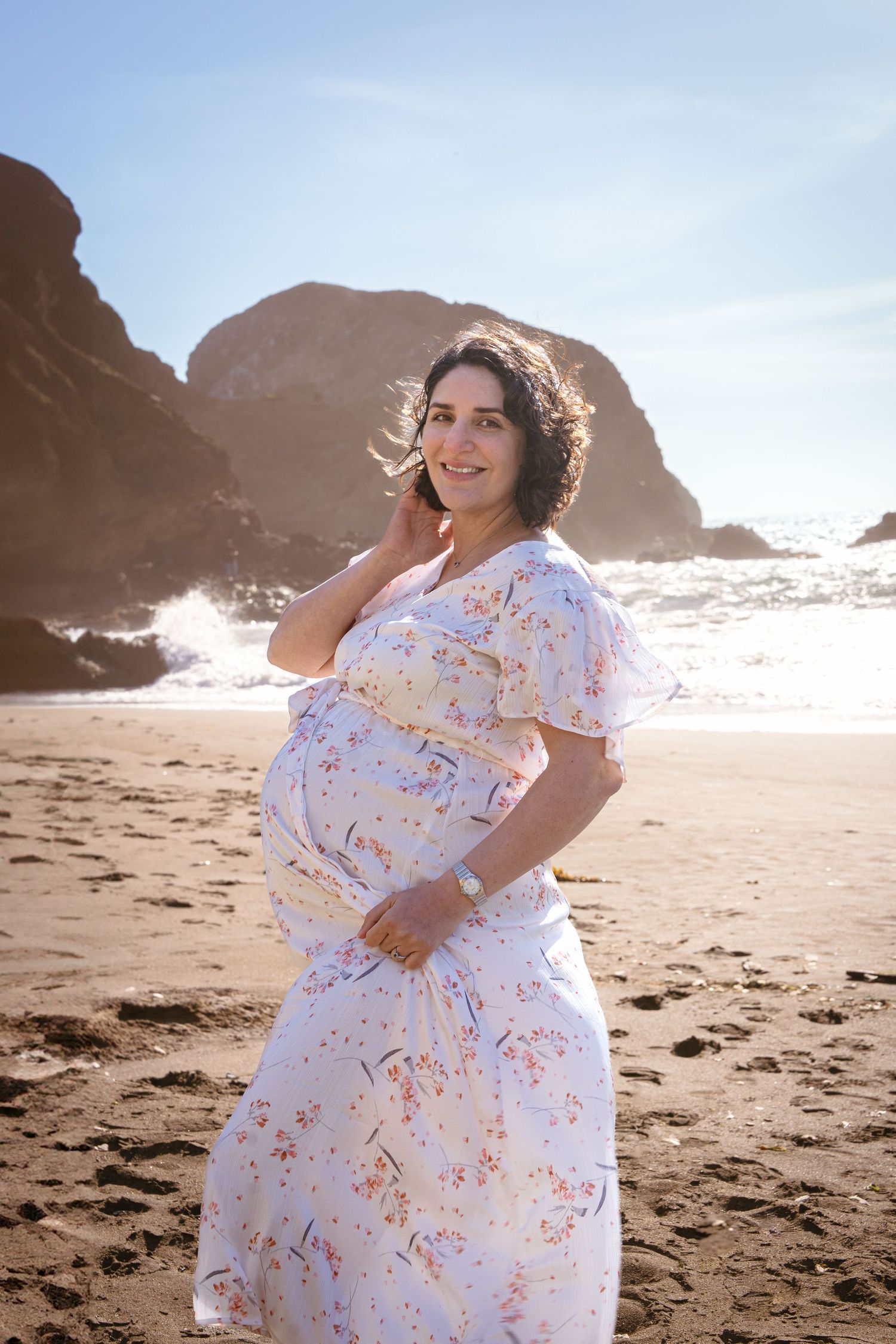 A pregnant person in a white floral dress poses on a beach with rocky cliffs in the background during sunset.