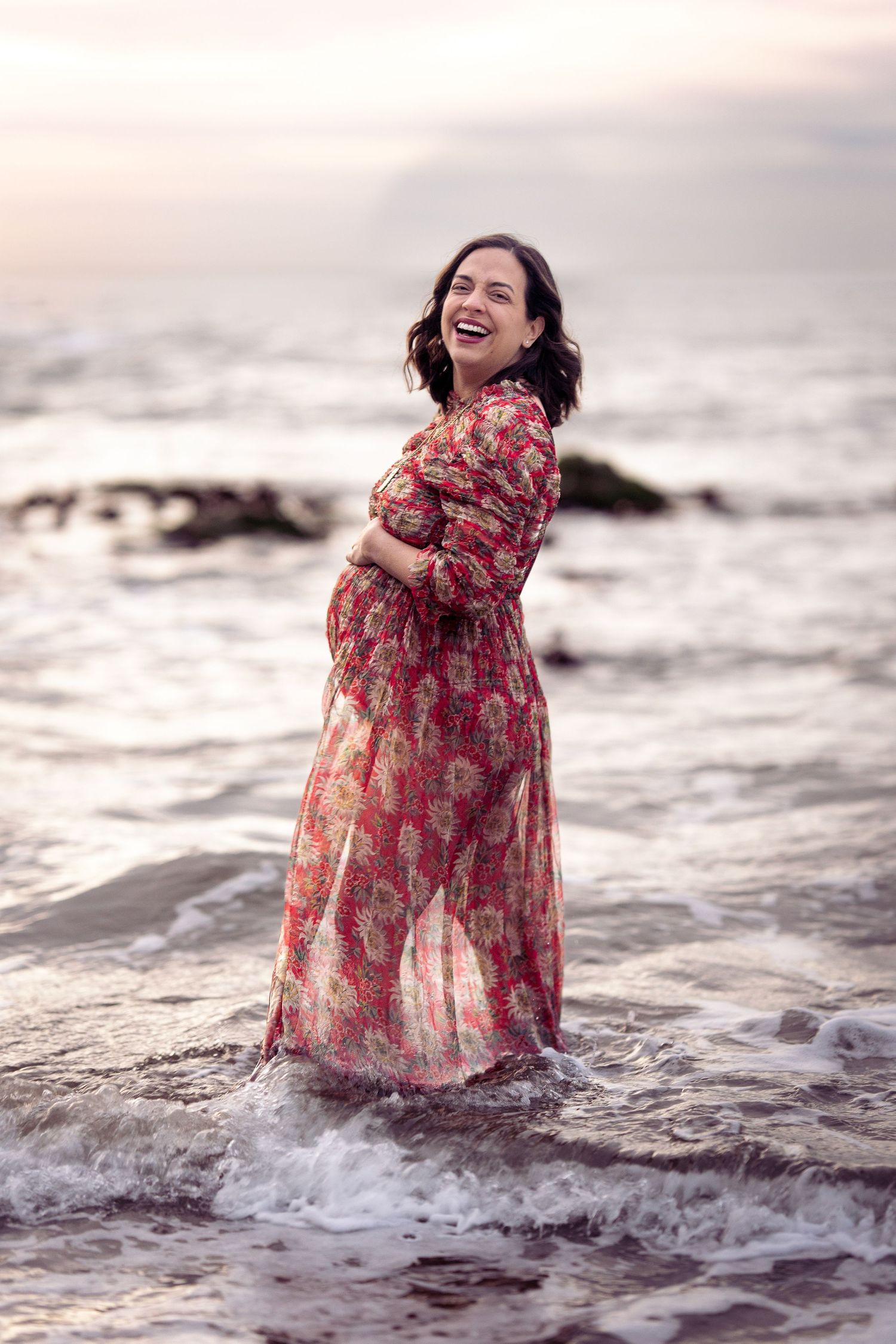 Expectant mother in flowing red dress stands in ocean waves at sunset.