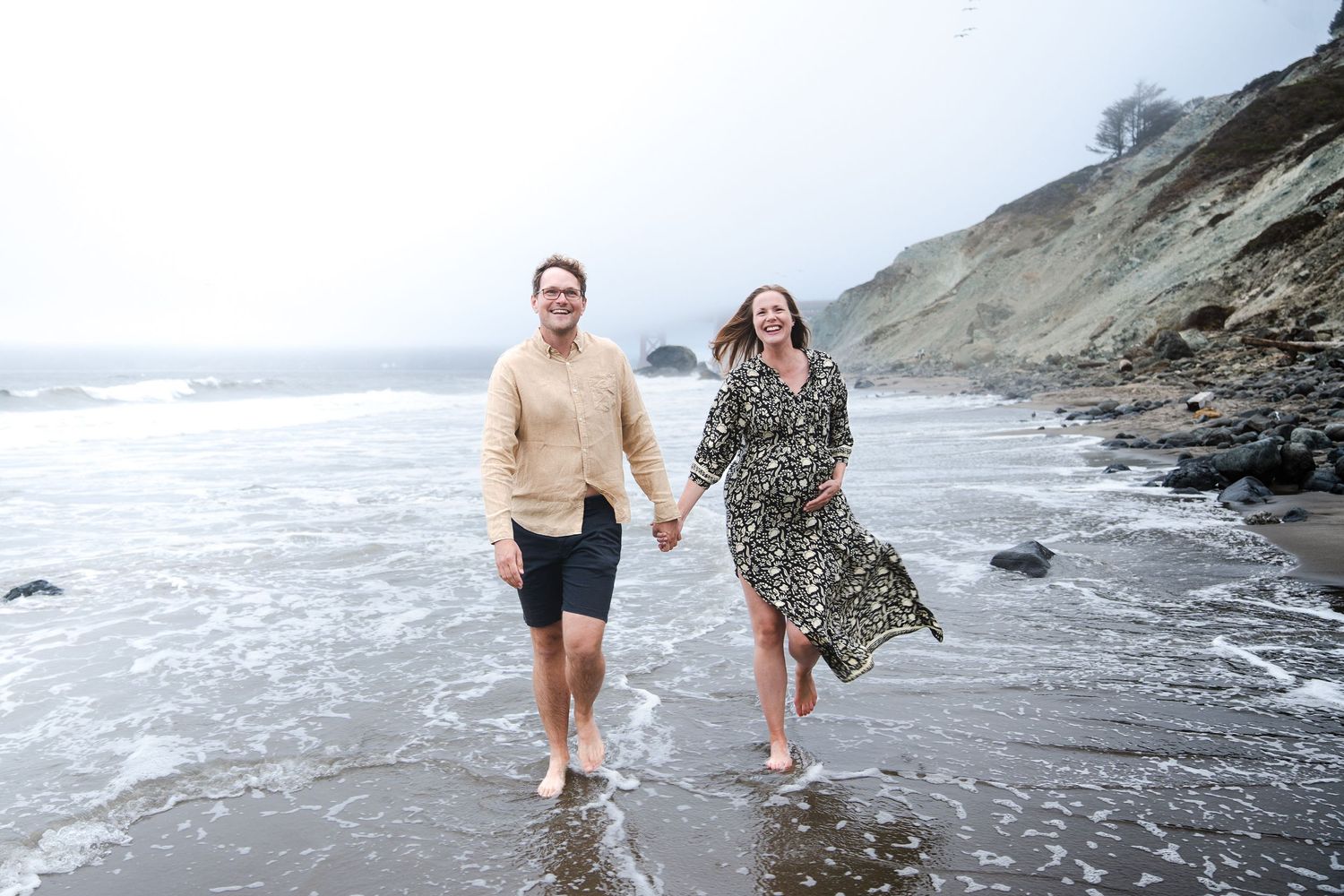 Couple running joyfully along a misty beach shoreline in casual attire.