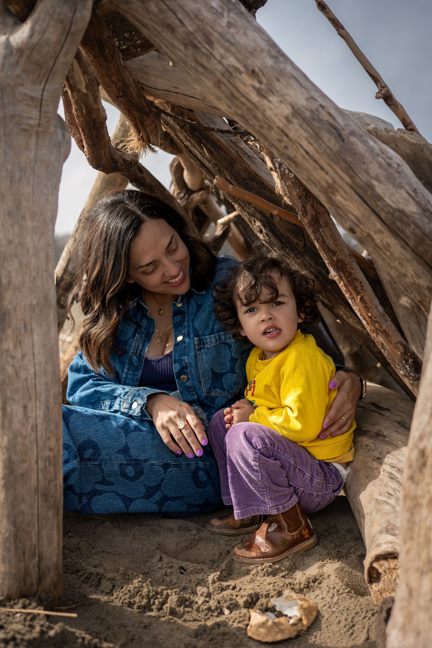A family takes shelter in a cozy driftwood beach fort on a sunny day, sharing a tender moment together.