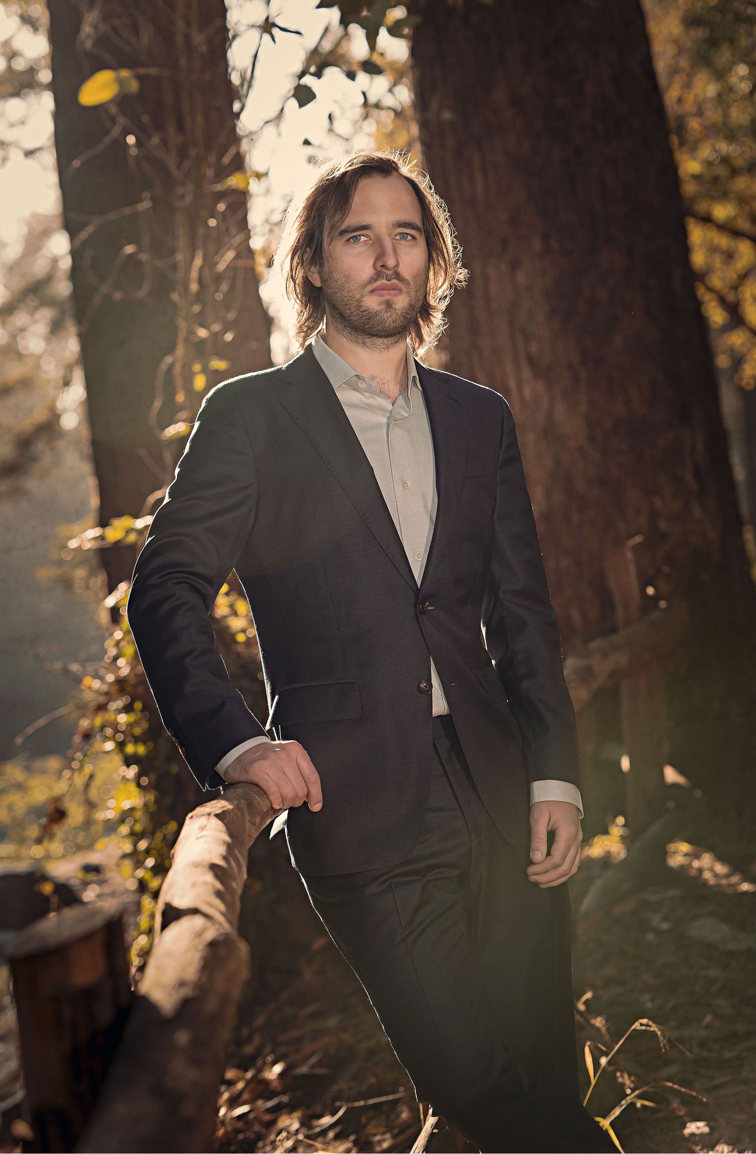 A person in a dark suit stands in dappled sunlight among autumn trees.